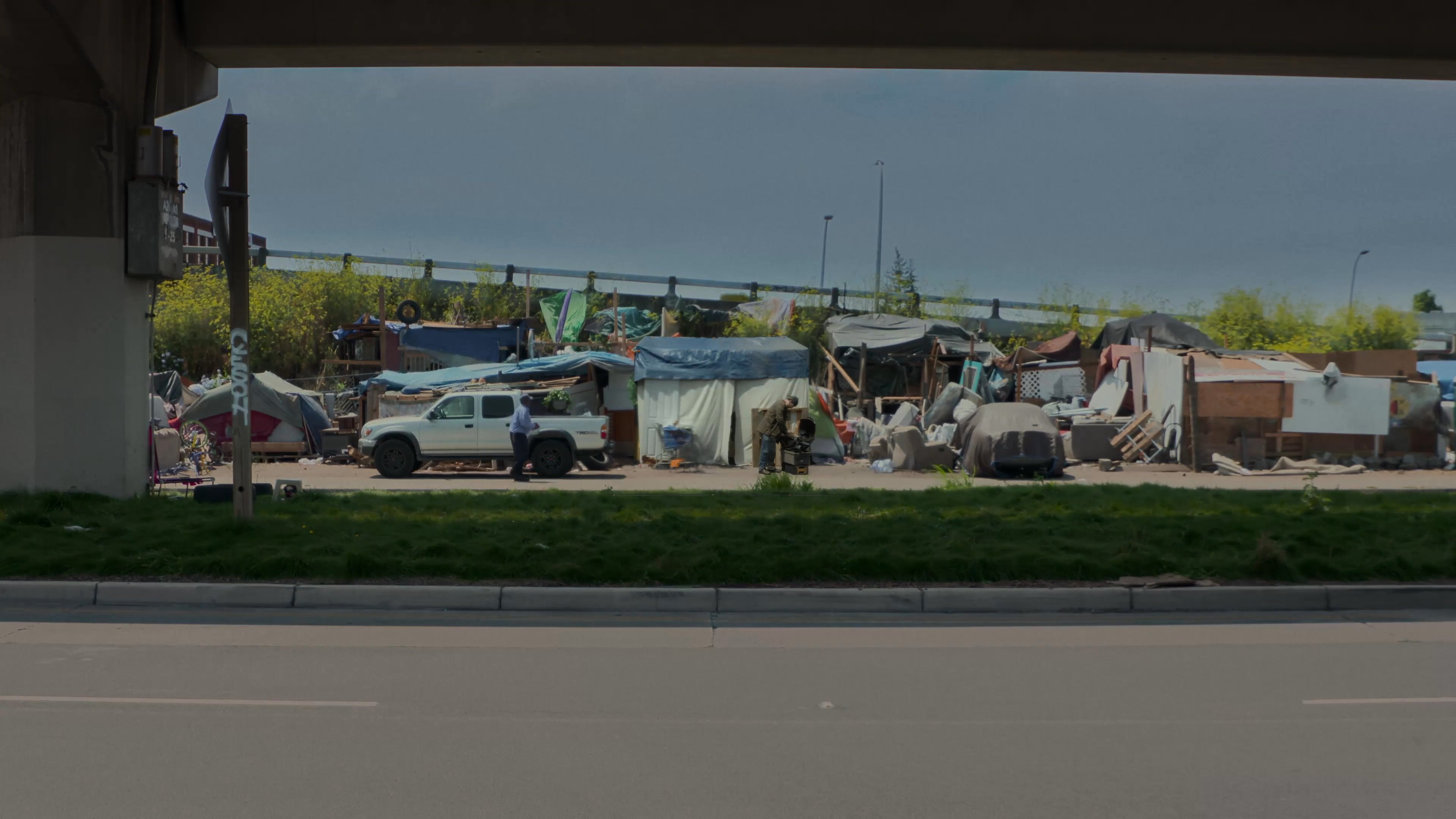 An urban landscape highlights a roadside homeless encampment consisting of multiple tents situated beneath a highway overpass in Minneapolis. The sequence captures the proximity of the temporary shelter to major transportation infrastructure.