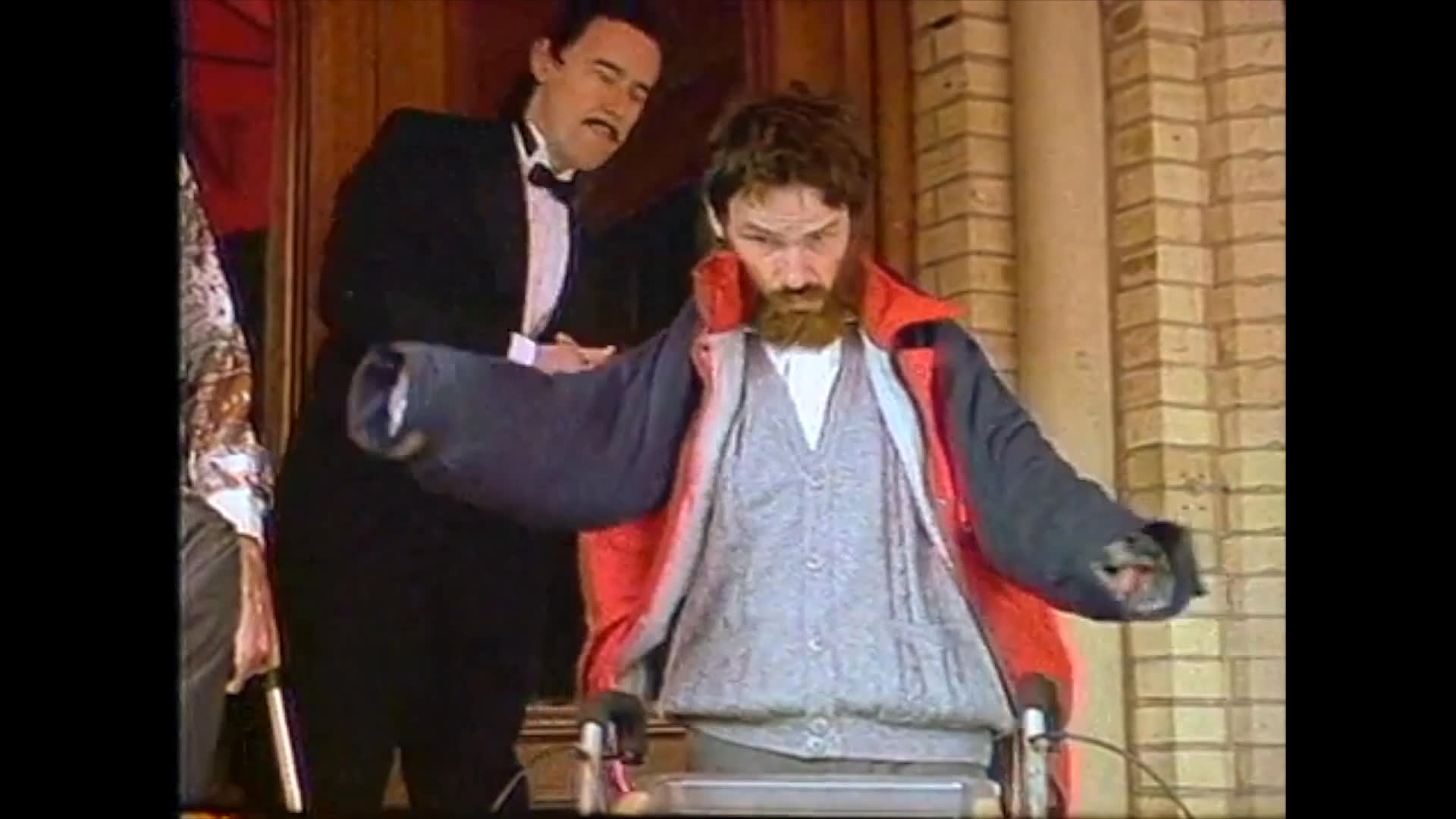 A man in a casual sweater and jacket struggles to put on his coat outside a theatre entrance while a man in a tuxedo watches nearby.
