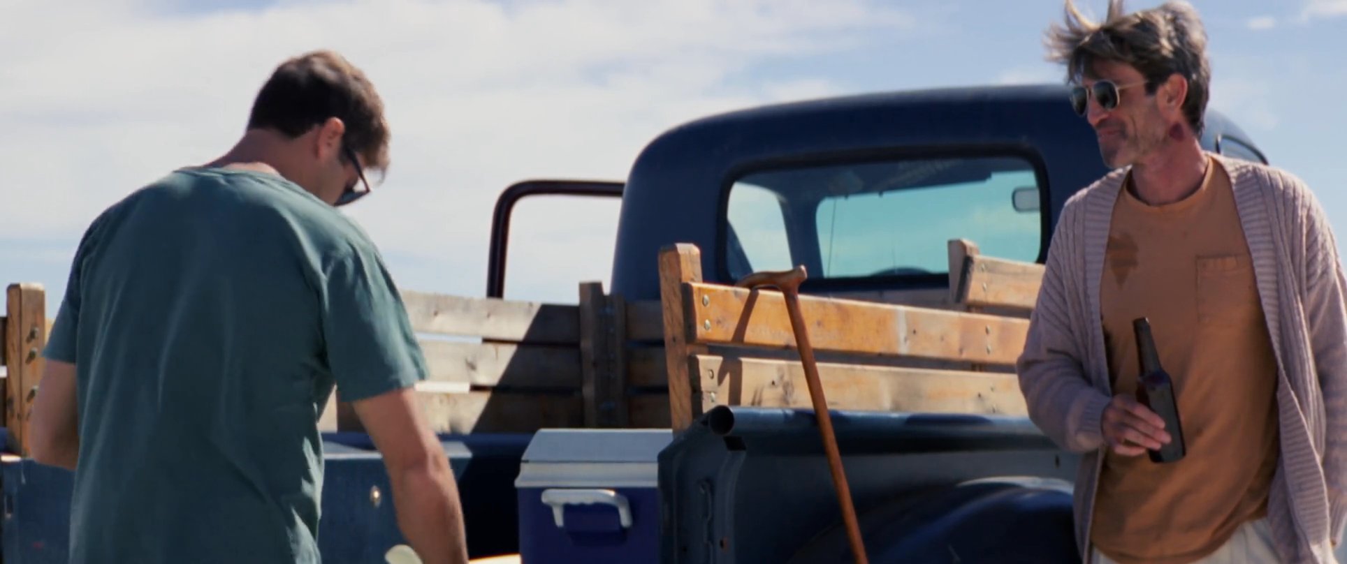 Two men enjoy a moment of relaxation by sharing cold beverages while leaning against a pickup truck in a sprawling desert landscape.