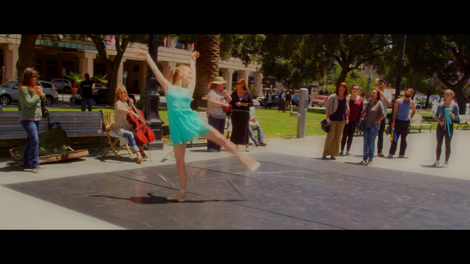 A ballet dancer performs a graceful routine on an outdoor stage in a park while a small audience watches. The performance features expressive movements and technical footwork in a public setting.
