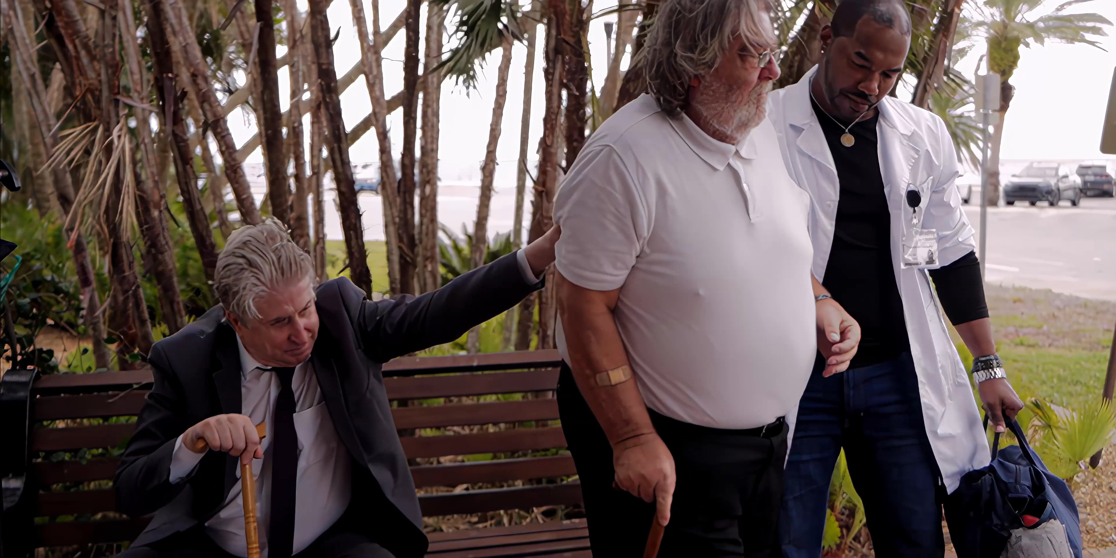 A caregiver in a medical coat assists an elderly man in rising from a park bench. Once the man departs, another individual sits down to continue the interaction with the gentleman remaining on the bench.