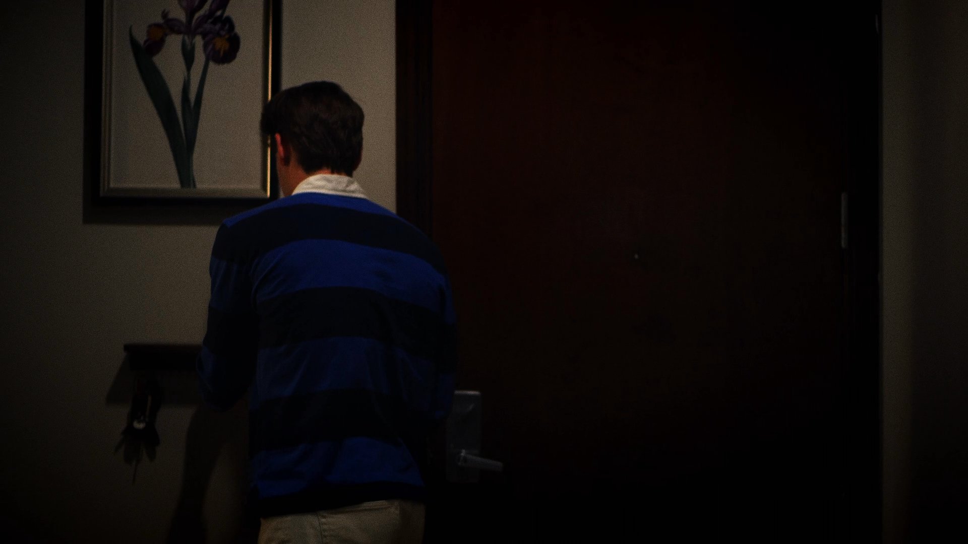 A woman observes through a door peephole as a man enters an apartment and begins searching through the refrigerator. The interaction suggests a tense, unexpected encounter within a residential space.