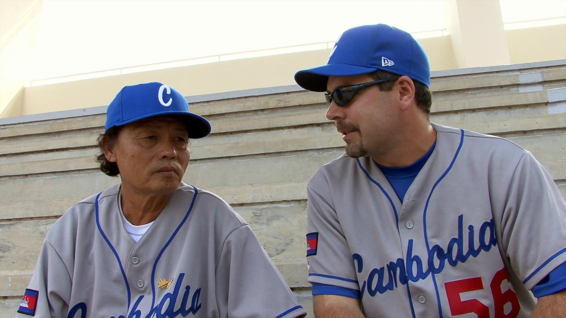 Two individuals in Cambodia baseball jerseys engage in an interview discussion while seated in a stadium seating area. The scene transitions to a wide shot of players on a baseball field.