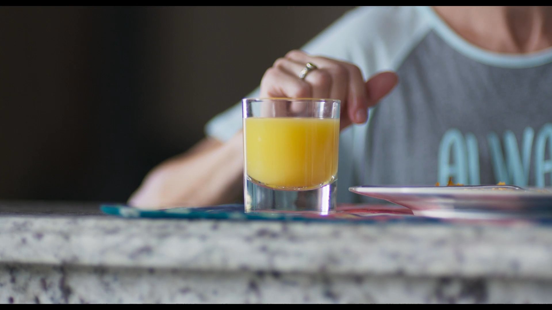 A woman sits at a breakfast table, rhythmically tapping her fingertips against a glass of orange juice while appearing lost in thought. The quiet morning scene captures a moment of pensive waiting.