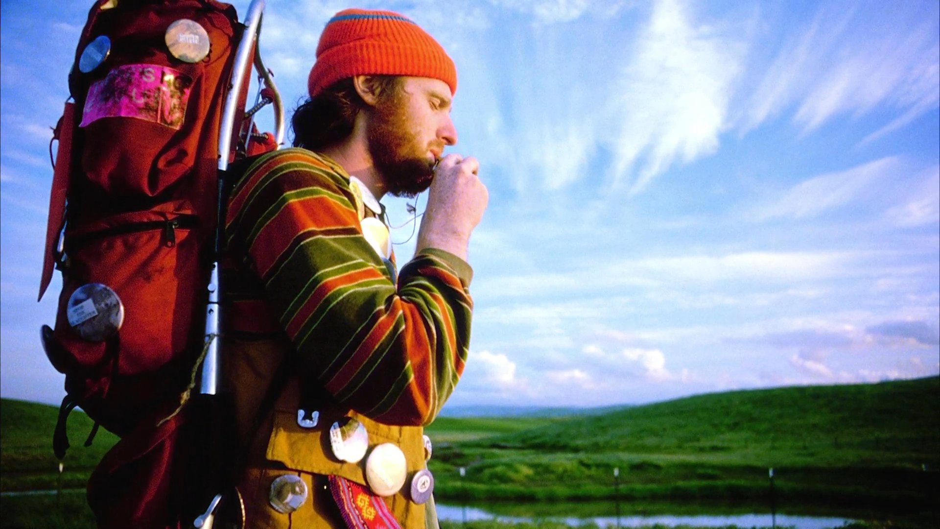 A bearded hiker wearing a red beanie and a heavily laden backpack pauses to inspect an item while standing in a vast, open landscape under a dramatic sky.