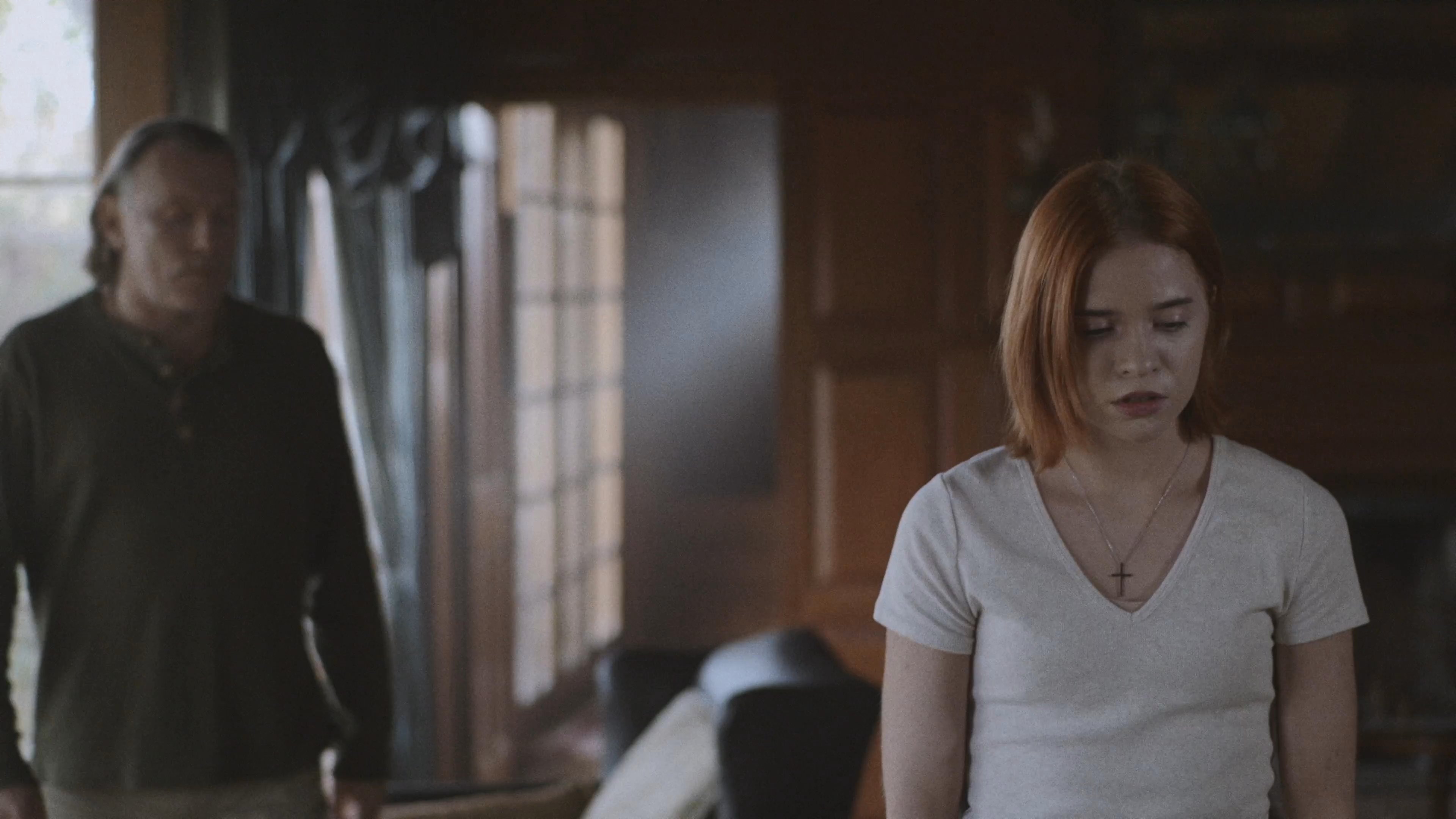 An older man approaches a younger woman standing in a dimly lit, rustic living room to initiate a somber conversation. The setting conveys a sense of isolation and dramatic tension between the two individuals.