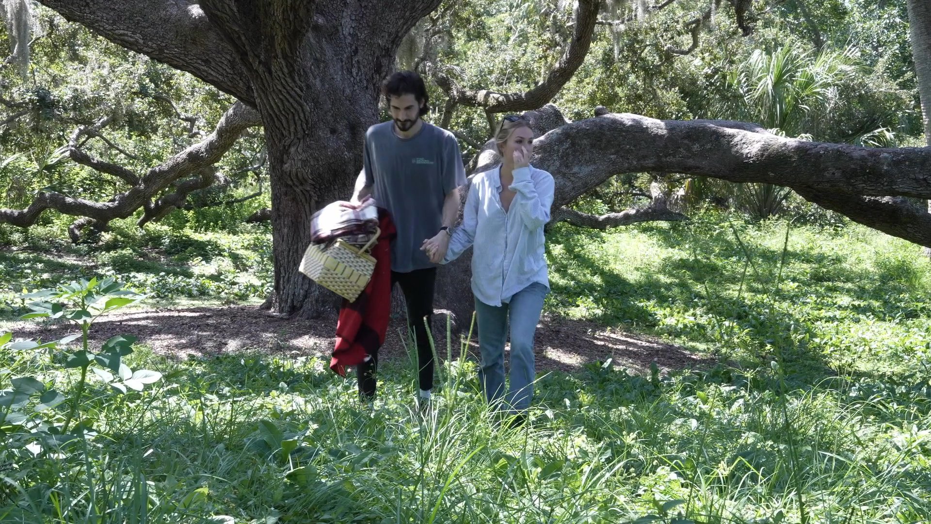 A couple walks through a grassy, tree-filled area while carrying a picnic basket and blanket before returning to their home.