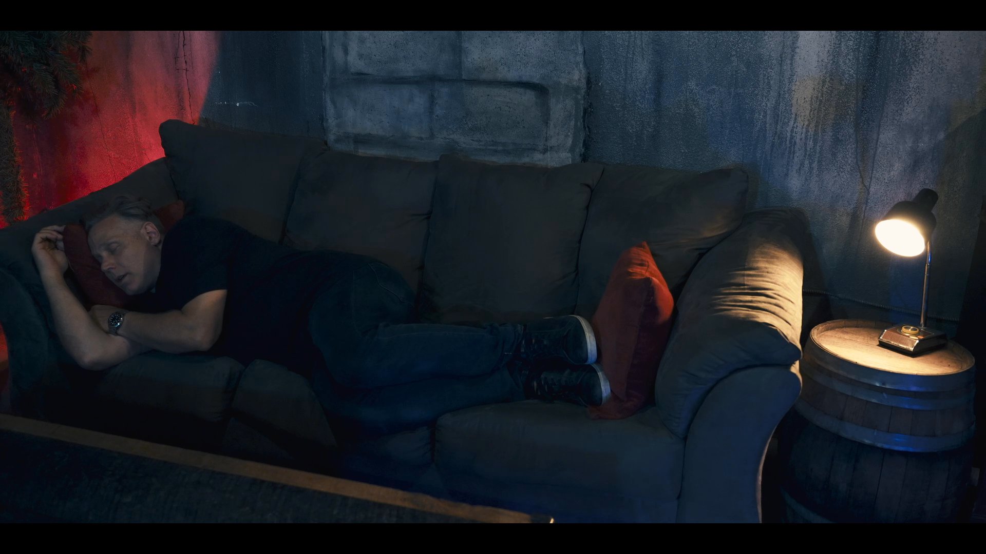 A man rests on a couch in a dimly lit room, framed by a prominent pink curtain. The scene shifts from his repose to a final shot of a modern, blue-lit interior space.