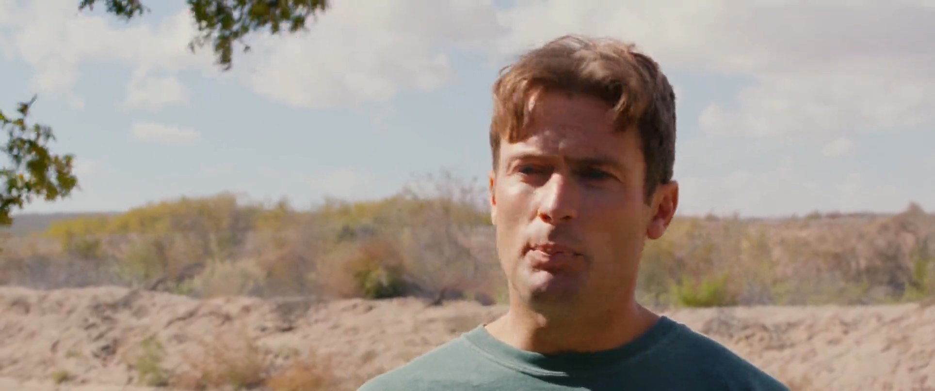 Two men engage in a conversation while standing outdoors in a natural, tree-lined environment. One individual wears sunglasses and a hooded garment, eventually removing his eyewear as they interact.