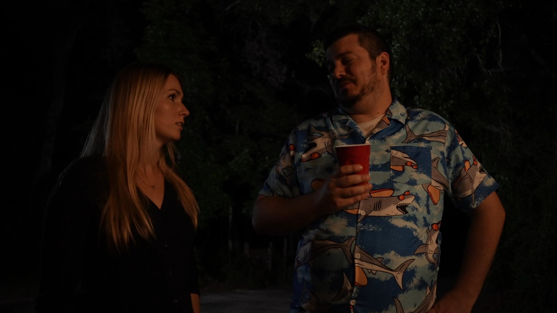 A man and a woman stand together outdoors at night, engaging in a quiet conversation while the man holds a red plastic cup.