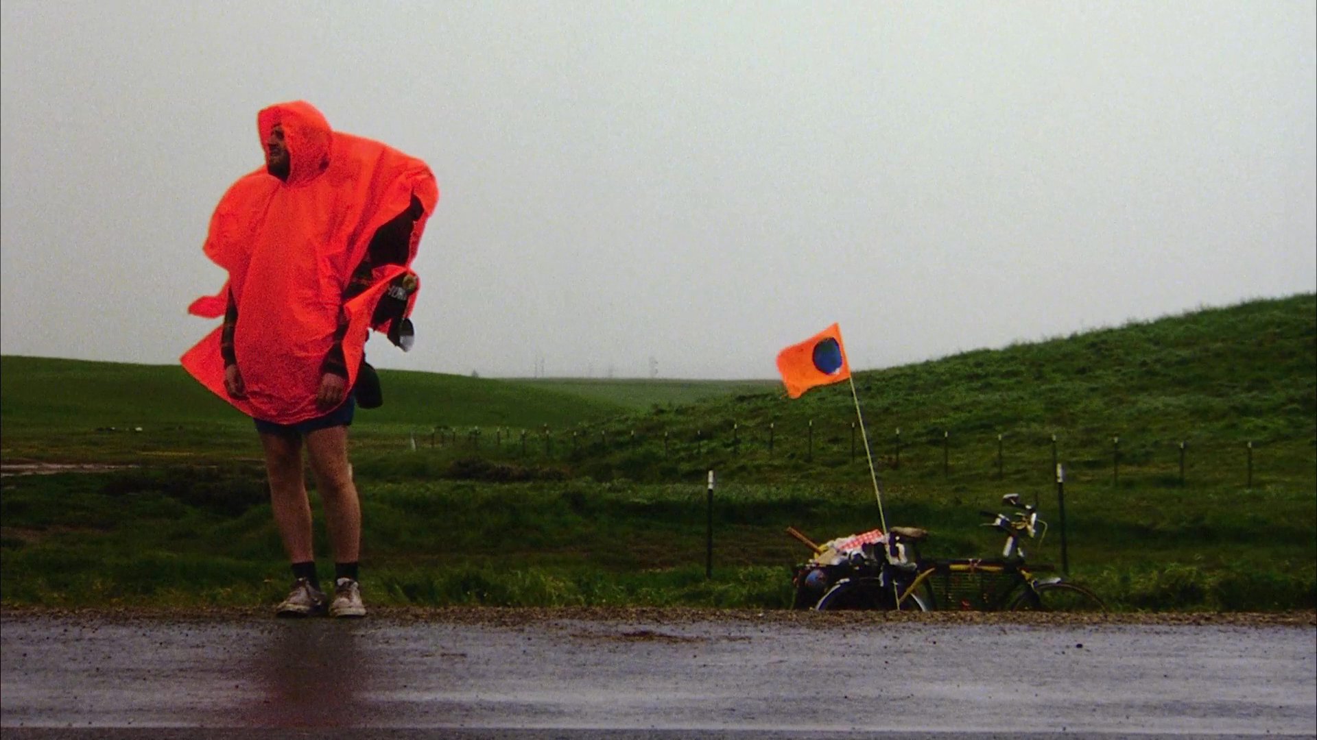 A long-distance cyclist rests near a field and later navigates a road while wearing a protective poncho during overcast conditions.