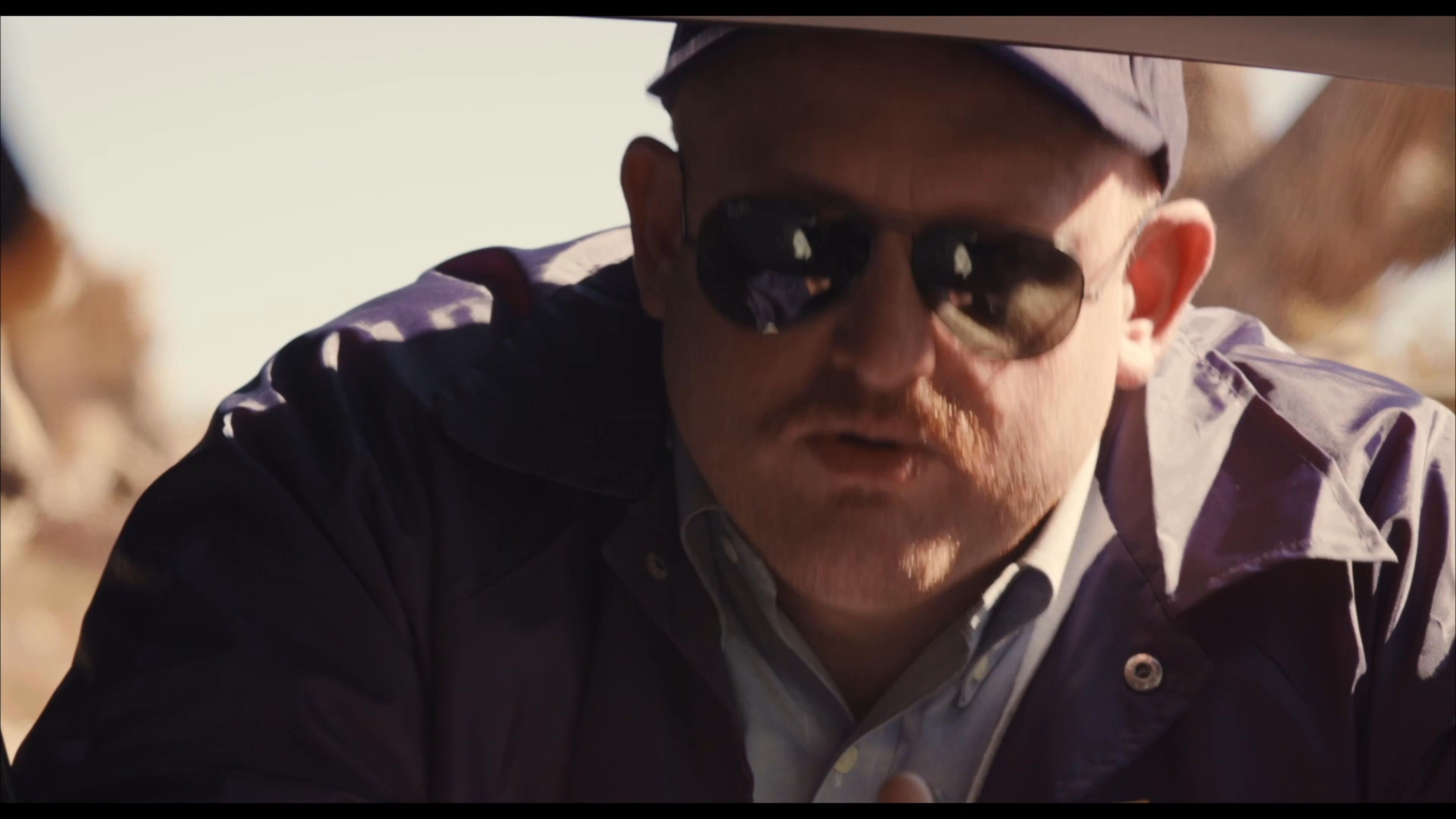 An investigator wearing a cap and sunglasses conducts a serious, authoritative interview while positioned outdoors in an arid, sunlit environment.