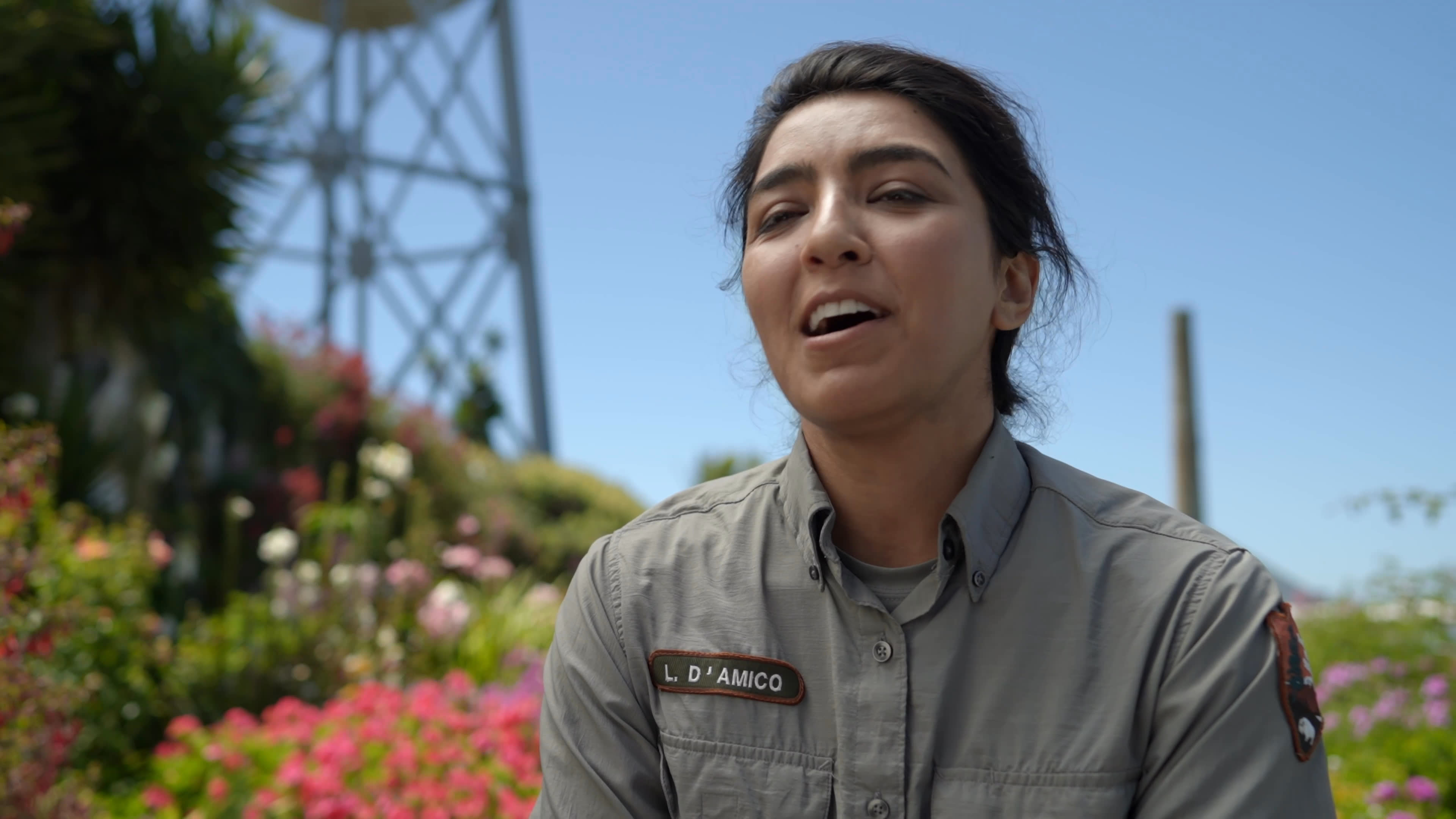 A uniformed worker speaks cheerfully about enjoying her bird-watching duties in a landscaped garden, while a falcon perches nearby. A metal tower rises behind the colorful flower beds.