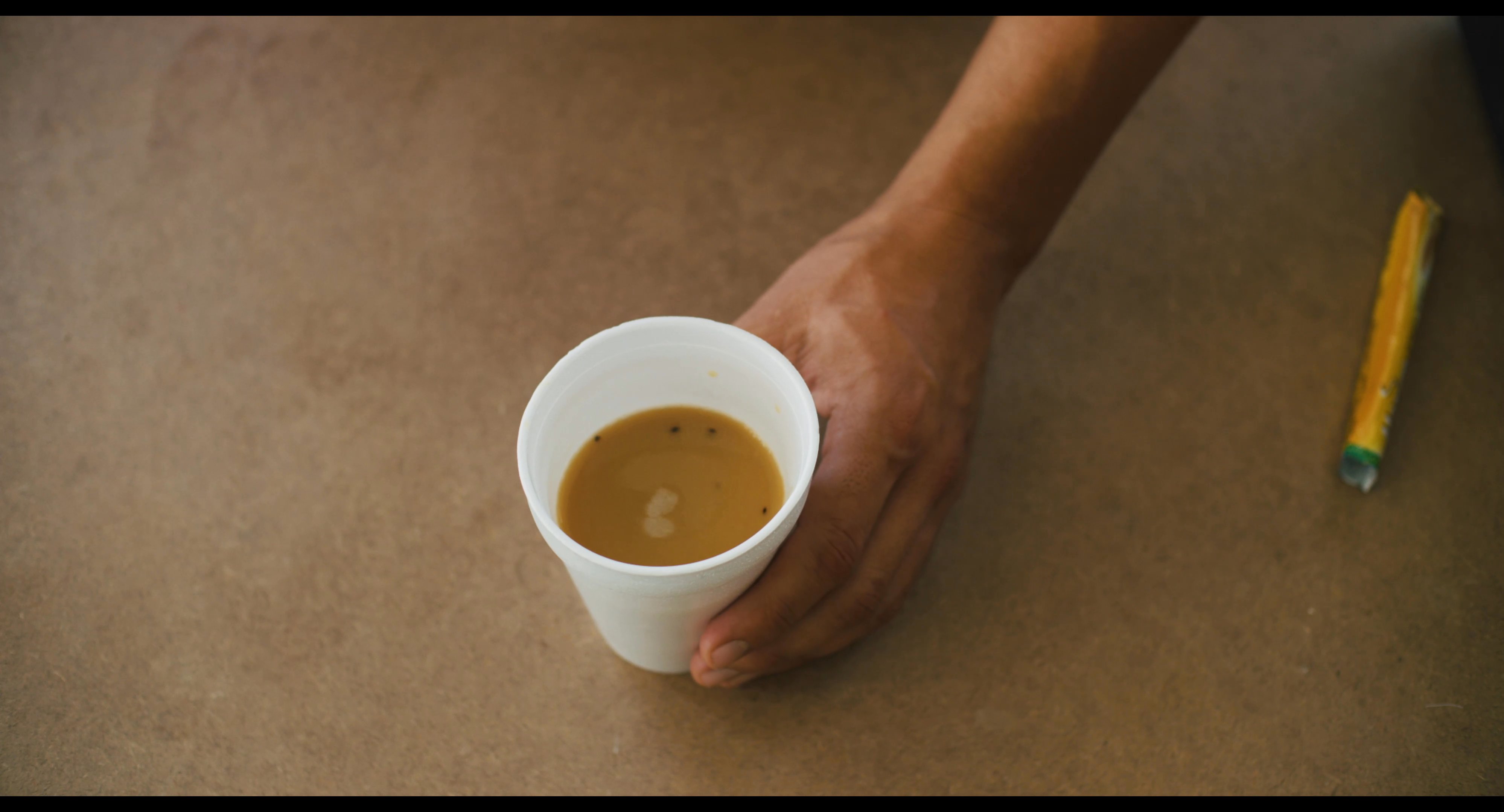 A woman prepares an instant coffee in a small shop while engaging in a casual conversation with a male colleague. The scene highlights a professional yet informal interaction within a retail environment.