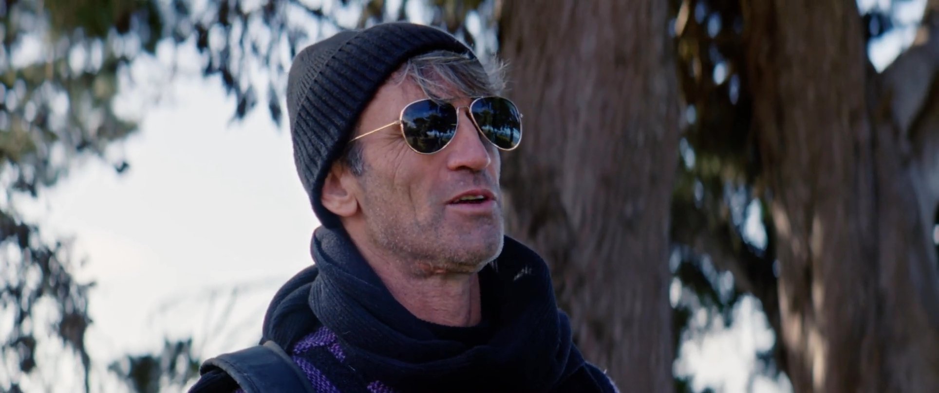 A man in a beanie and sunglasses engages in a conversation near the coast before the scene transitions to two other men wearing wetsuits.