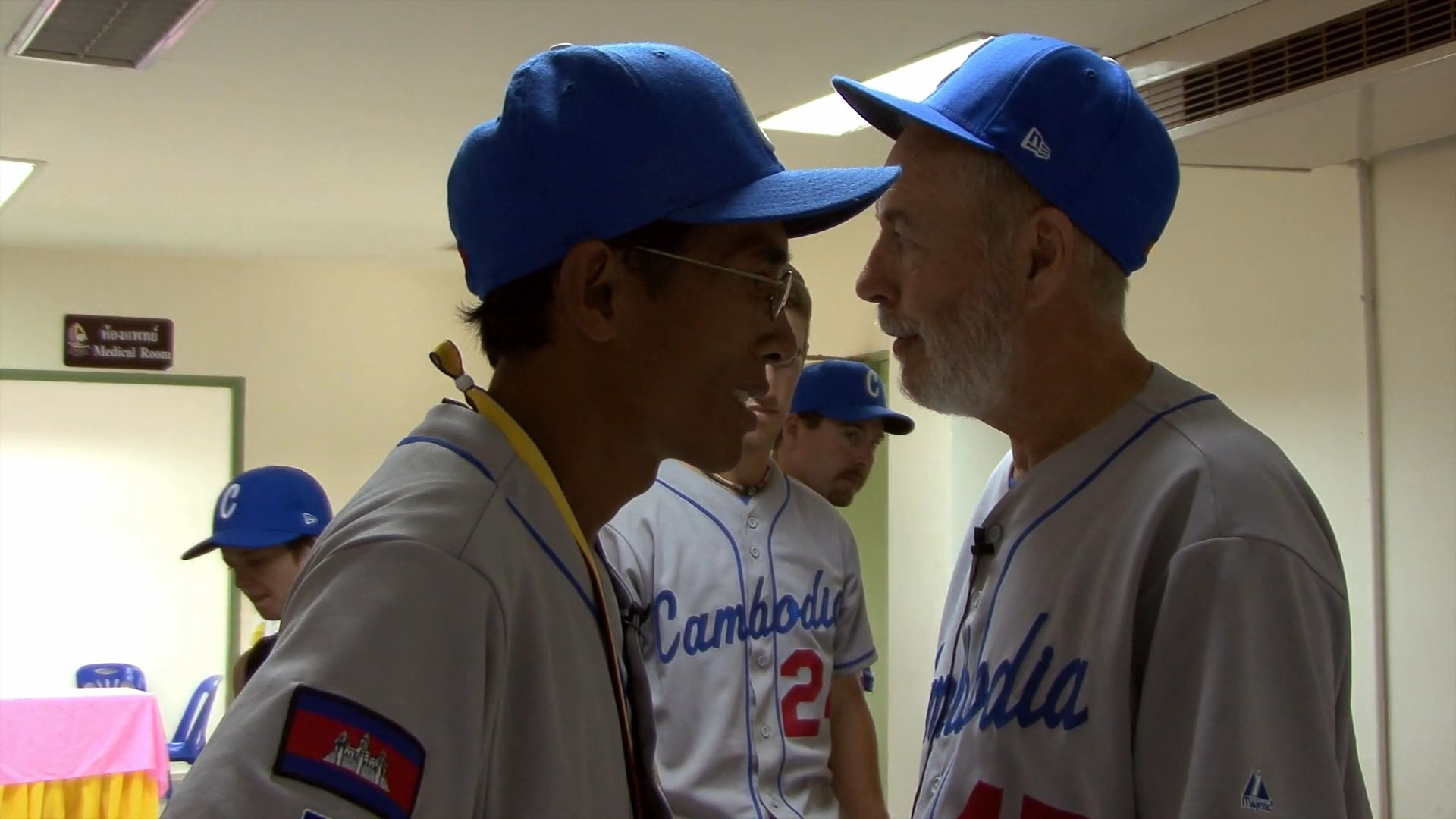 A coach and a baseball player in team uniforms engage in a focused conversation within a locker room environment. The pair discusses strategy while surrounded by other team members preparing for an event.