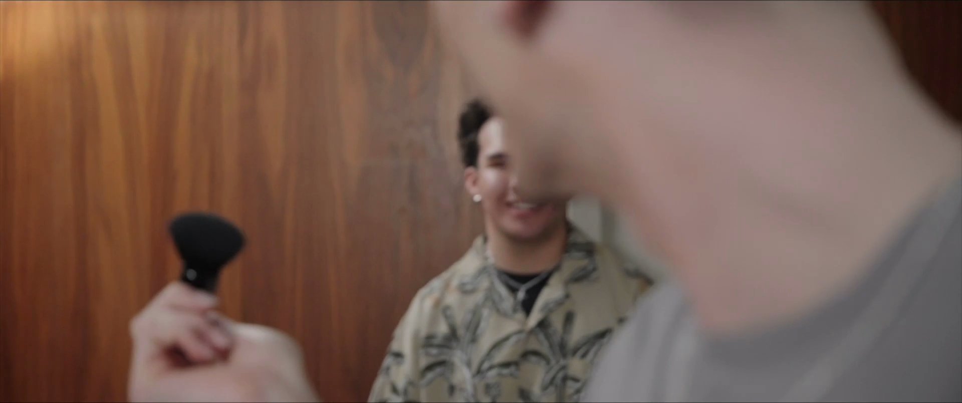 Two individuals interact in an indoor setting while one applies makeup with a cosmetic brush in a mirror. The sequence highlights personal grooming and preparation.