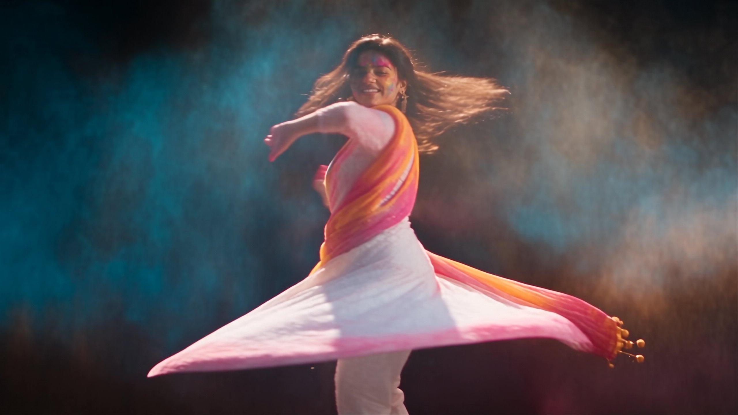 A dancer in a white and pink anarkali whips bursts of Holi powder across a black studio in widescreen.