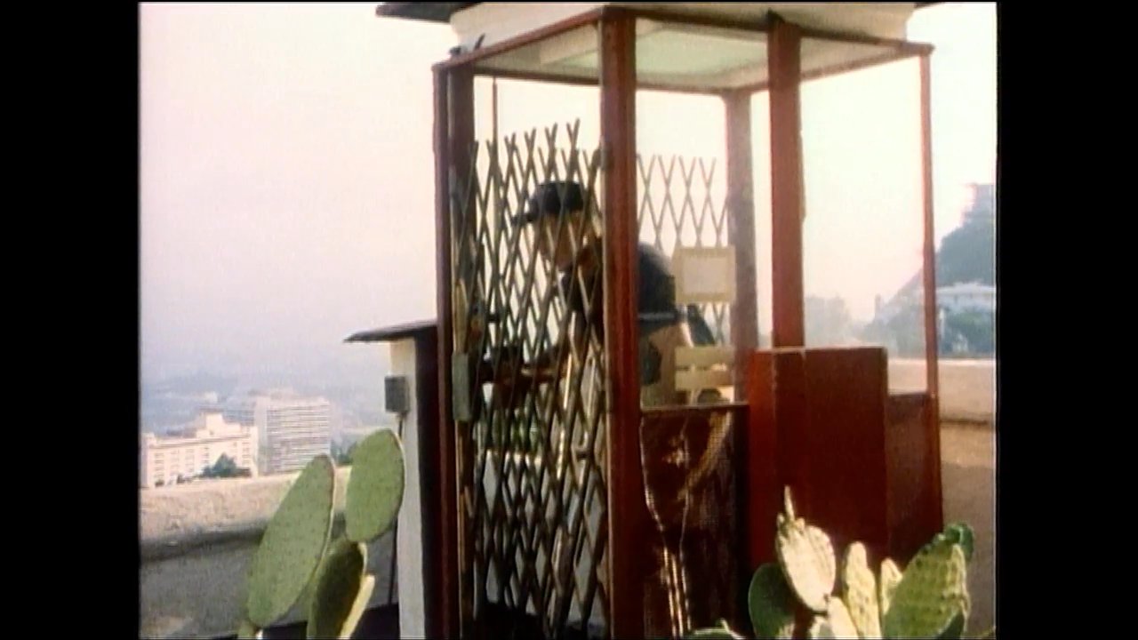 A man and woman converse on an elevated terrace overlooking a city, while a man using crutches exits an outdoor elevator and approaches a group of people nearby.