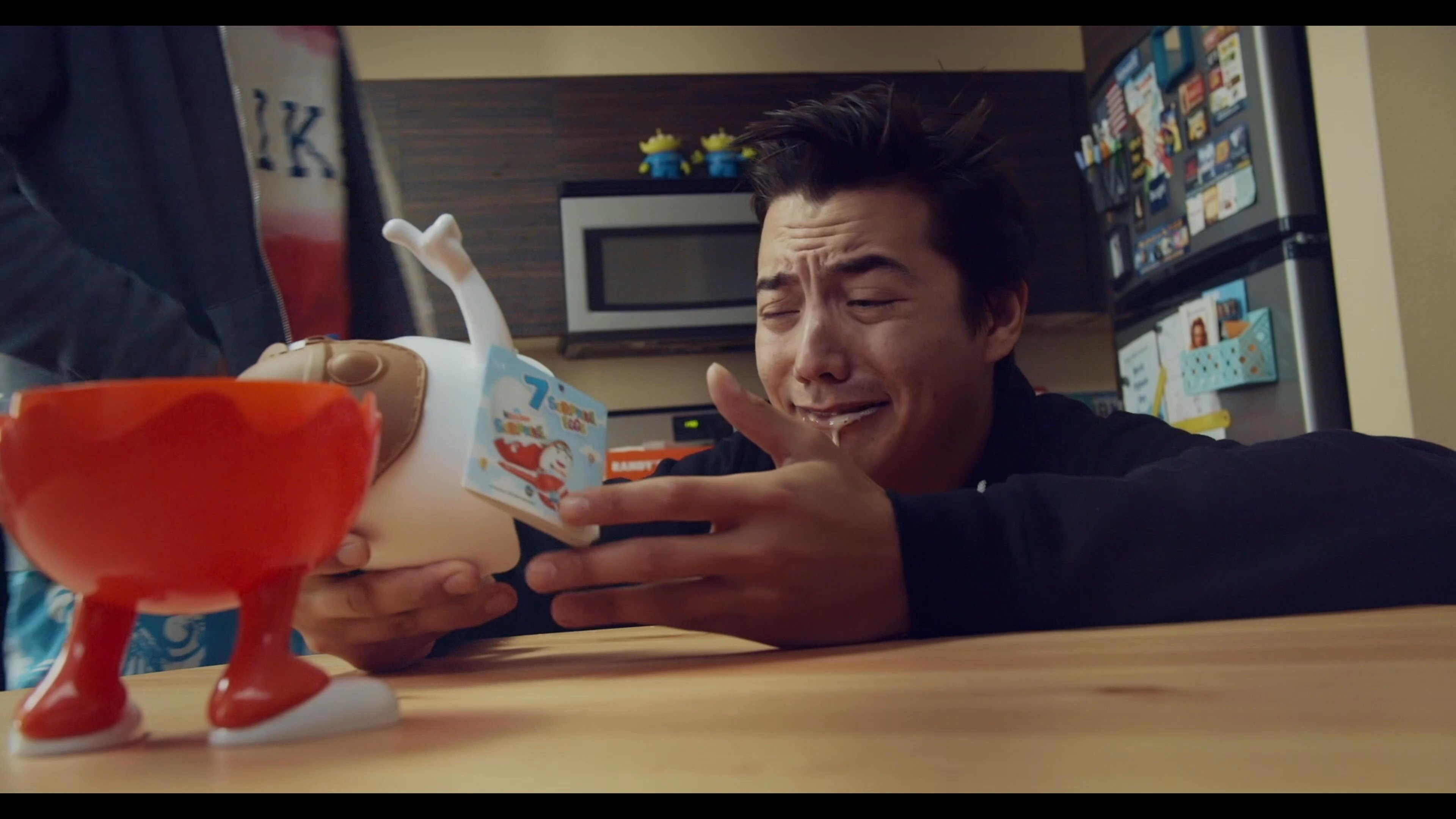 A man experiences a sentimental reaction while examining a small toy at a kitchen table. His friend observes from the living room, capturing a moment of personal reflection.