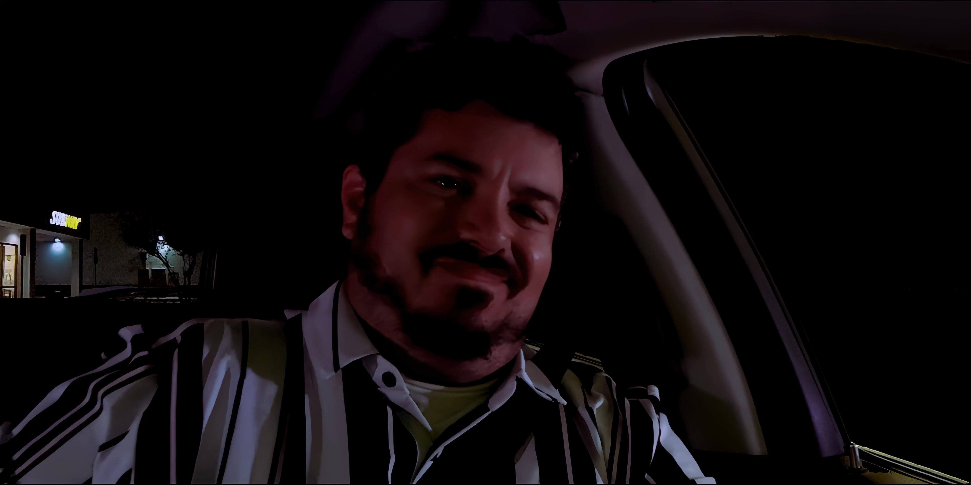 A man drives his vehicle at night and pulls into a parking lot, subsequently exiting the car to approach a storefront.