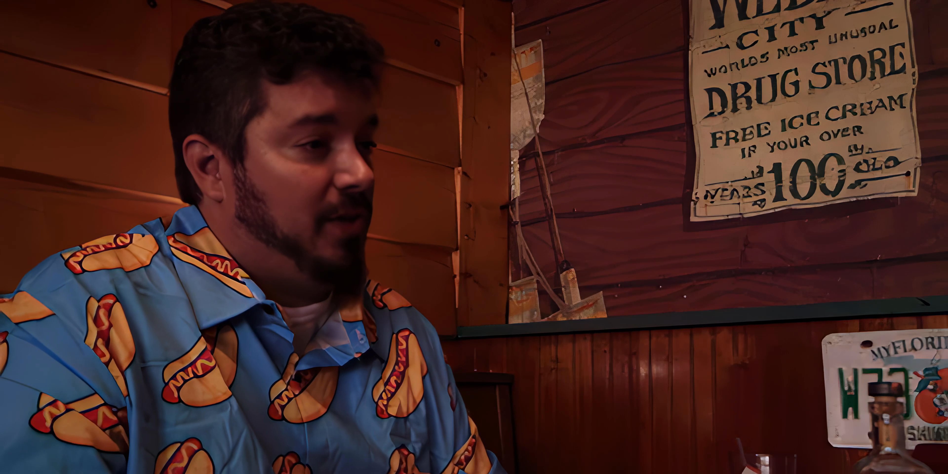 Two men engage in a focused discussion while seated inside a diner decorated with vintage advertisements. One man wears a formal suit while the other is dressed in a patterned shirt, as they exchange remarks in the booth.