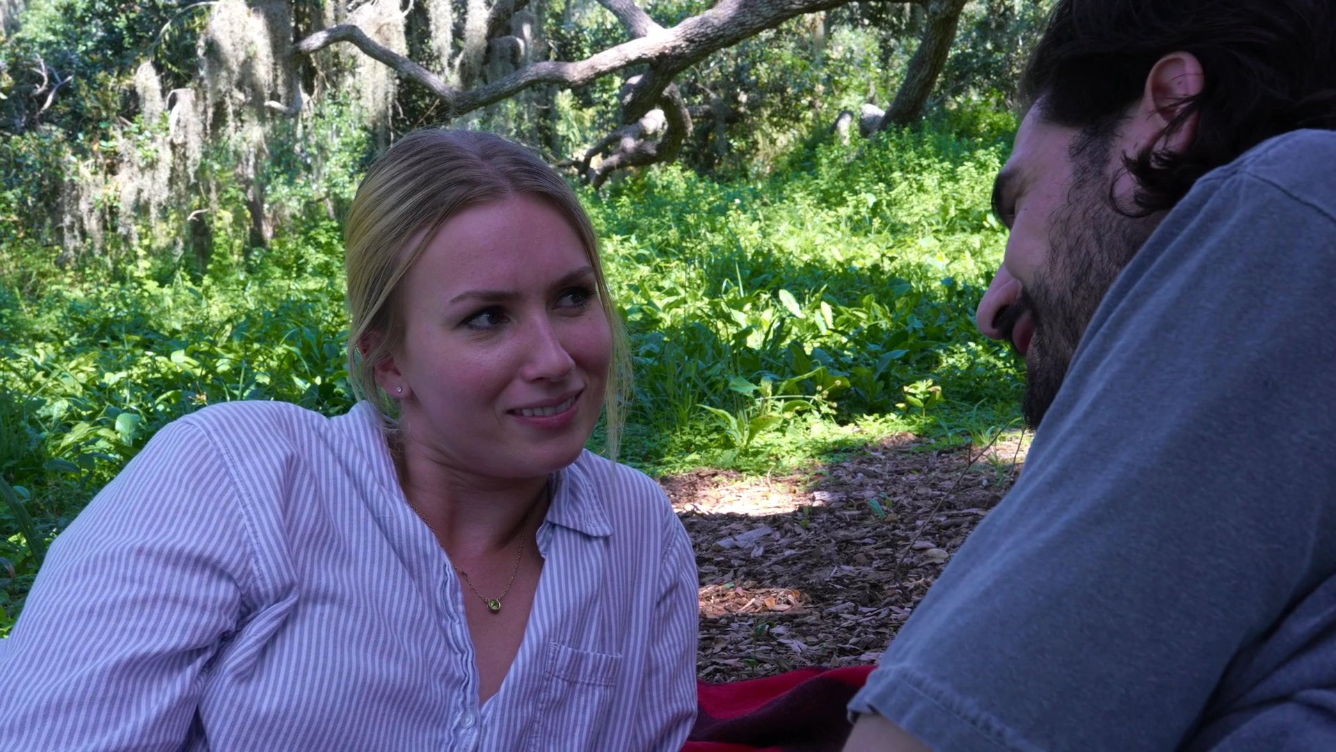 A man and woman engage in a lighthearted conversation while resting together on a blanket in a sunlit, wooded area. Their relaxed posture and warm smiles reflect a casual and pleasant interaction.