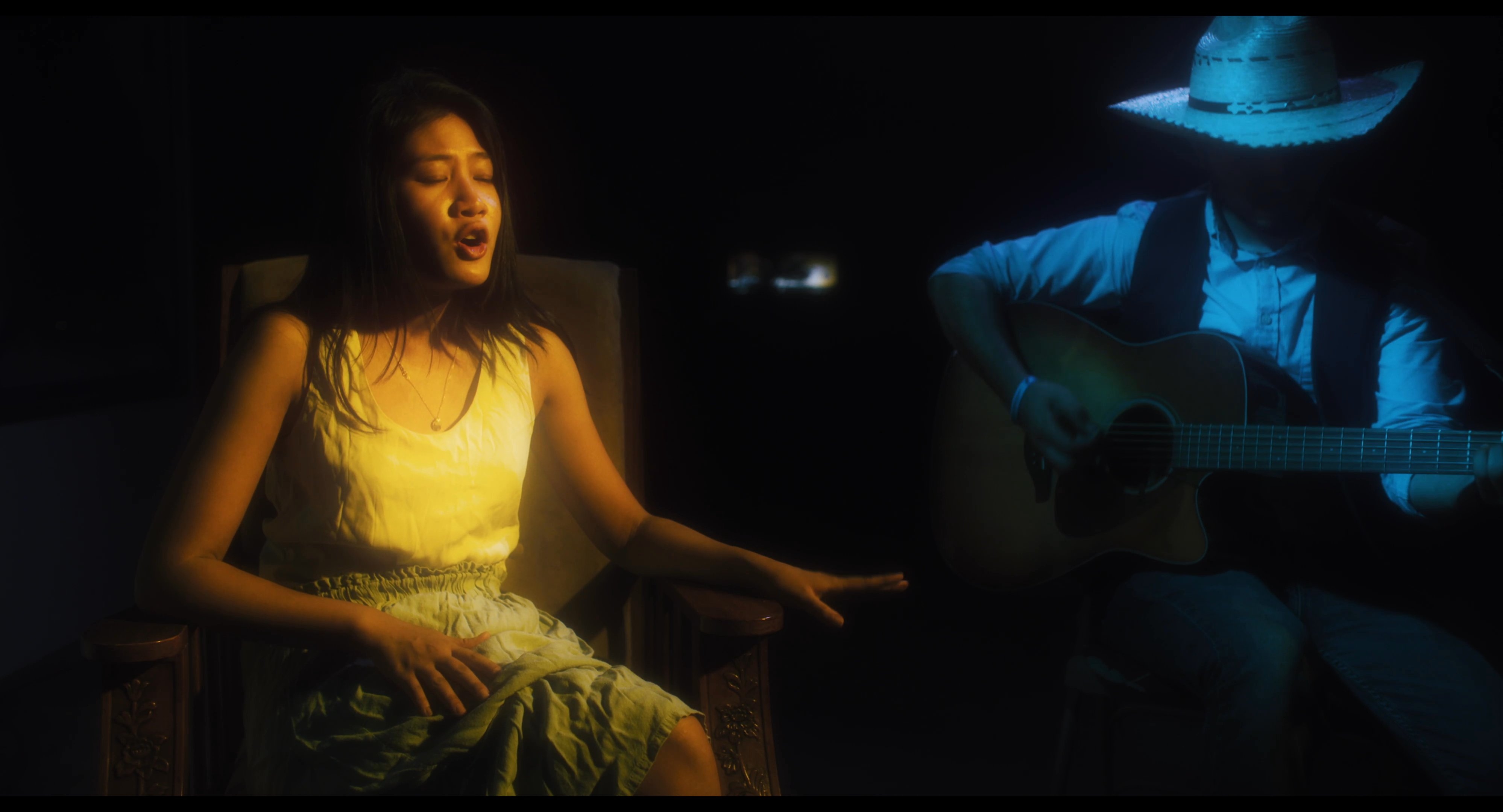 A vocalist performs while a guitarist wearing a cowboy hat accompanies her on acoustic guitar in a dimly lit setting. The duo focuses on their musical arrangement under dramatic stage lighting.