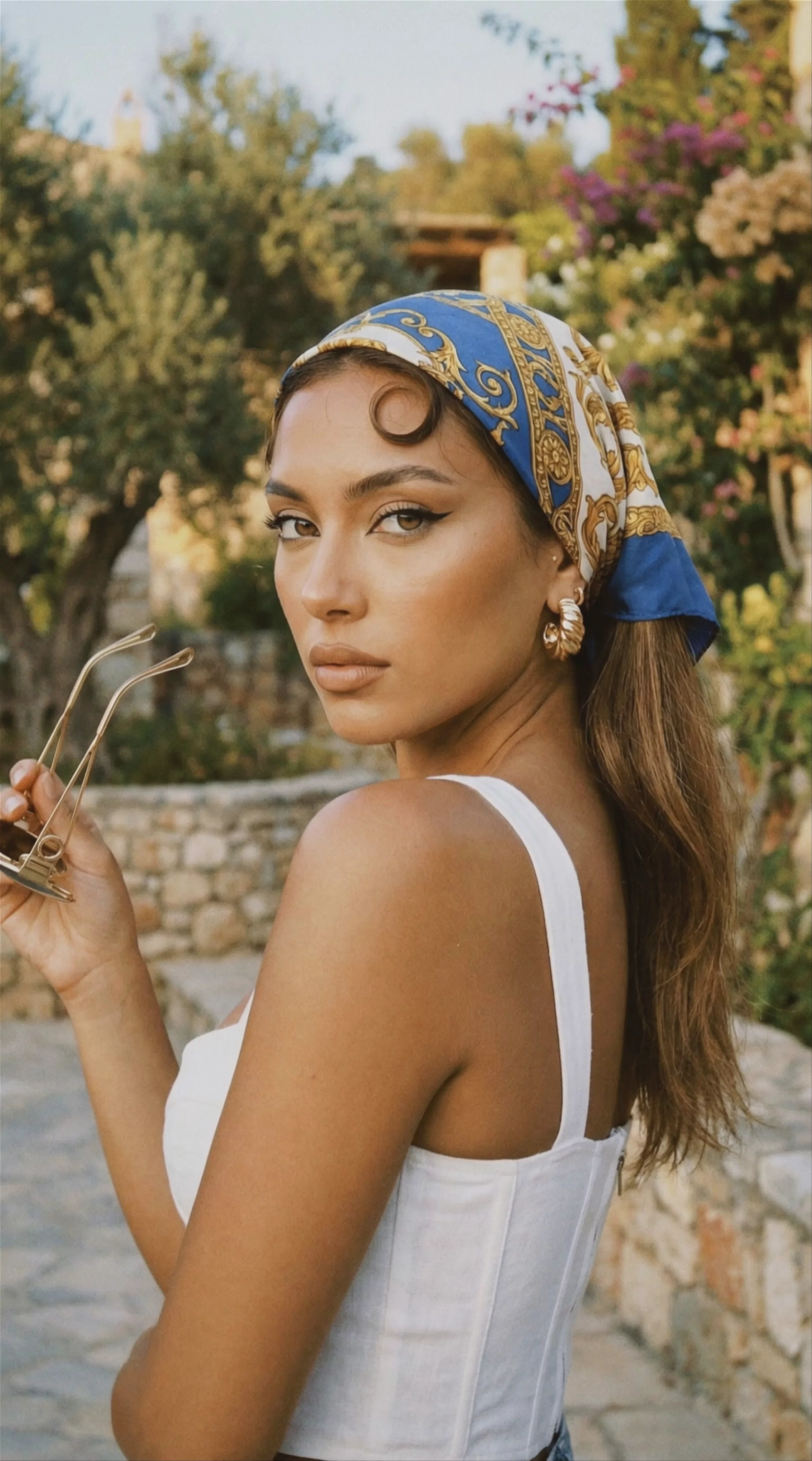 A vertical 1072x1928 video frame shows a young woman looking over her shoulder. She wears a blue, white, and gold patterned headscarf, a white top, and gold hoop earrings while holding sunglasses. Generated using Kling O3 Pro, the shot features a warm, sunlit outdoor background with stone walls and foliage.