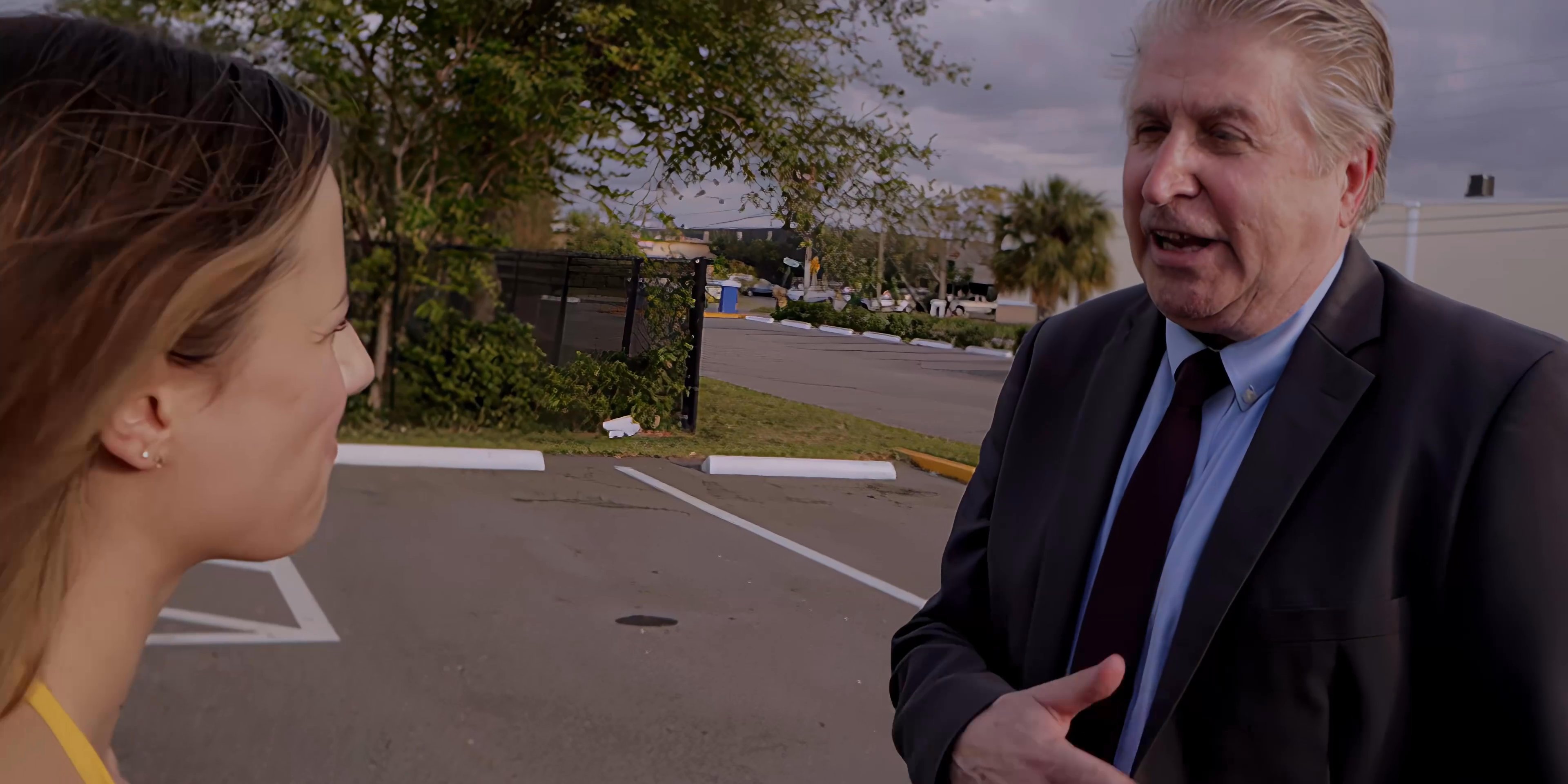 A man in a suit engages in a tense conversation with a woman dressed in a gold top in a parking lot. The exchange reflects a serious interpersonal or business-related interaction.