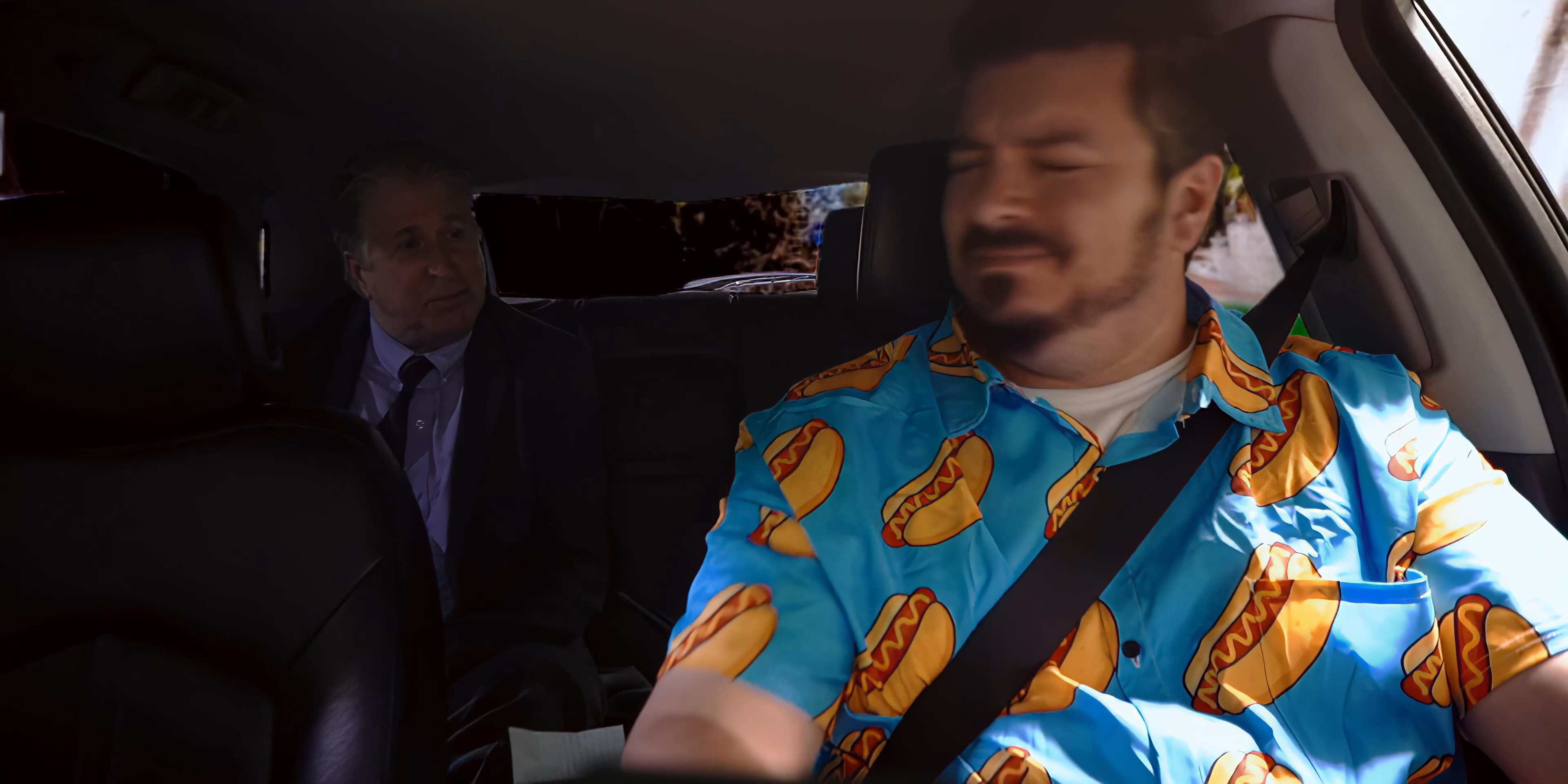 An older man in a suit engages in a discussion with a younger passenger wearing a vibrant patterned shirt while traveling in the backseat of a car.
