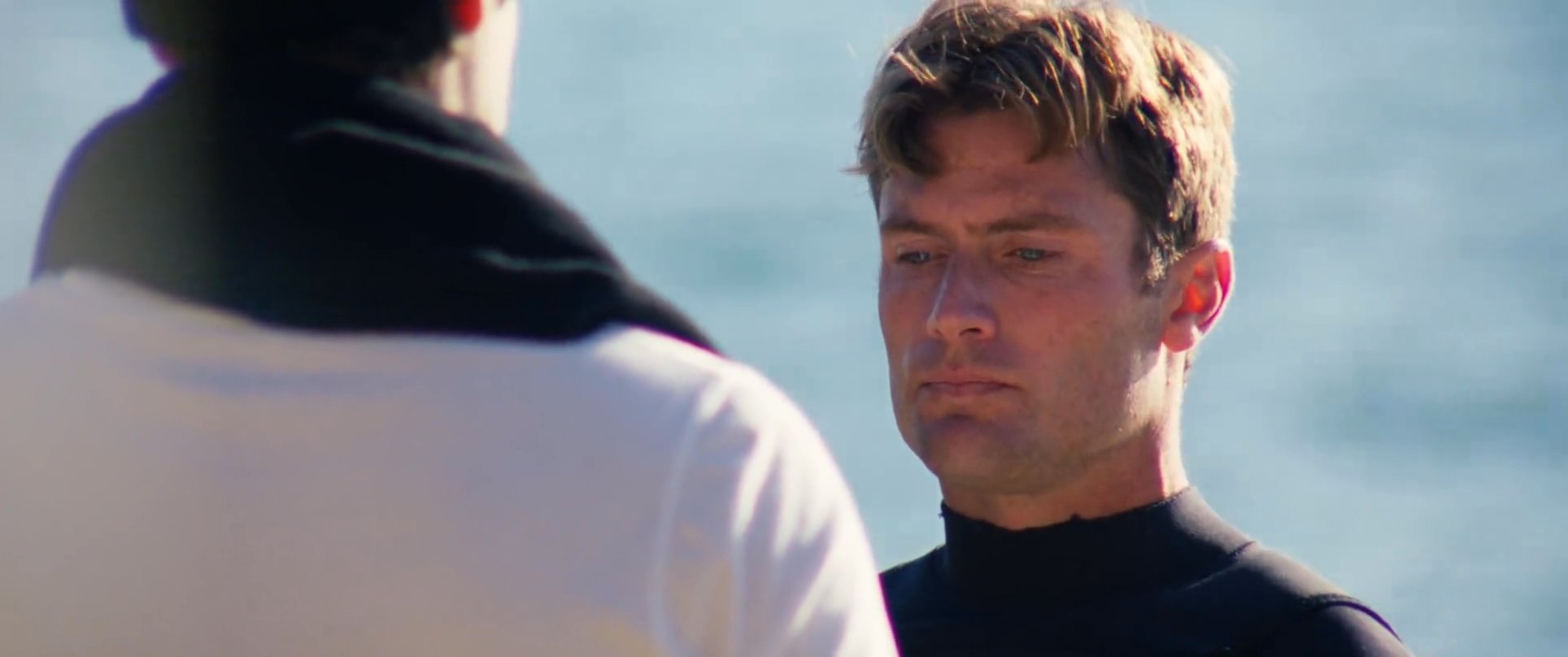 Two men engage in an earnest conversation against a coastal backdrop, concluding the interaction with a heartfelt embrace.
