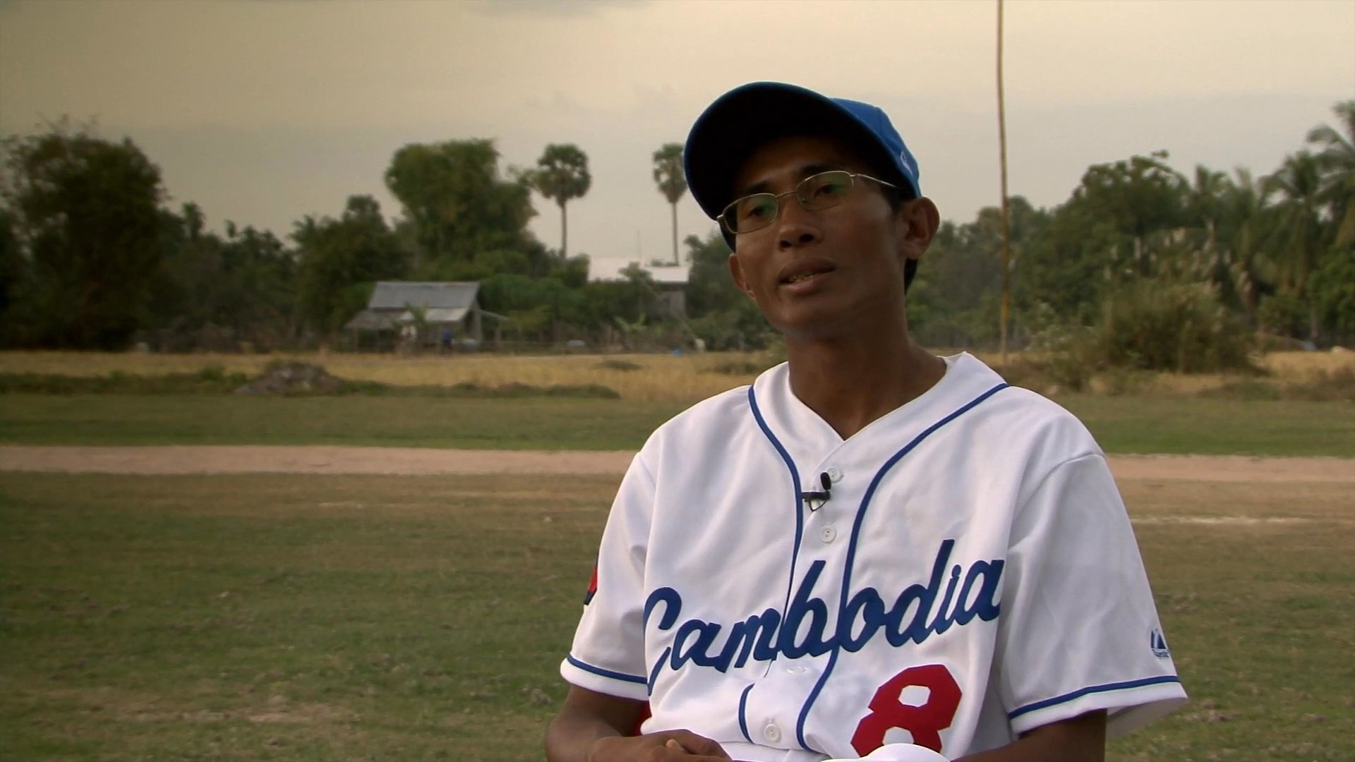 Footage transitions from a bustling outdoor market with local vendors to an interview with an athlete wearing a Cambodian baseball jersey on a sports field.