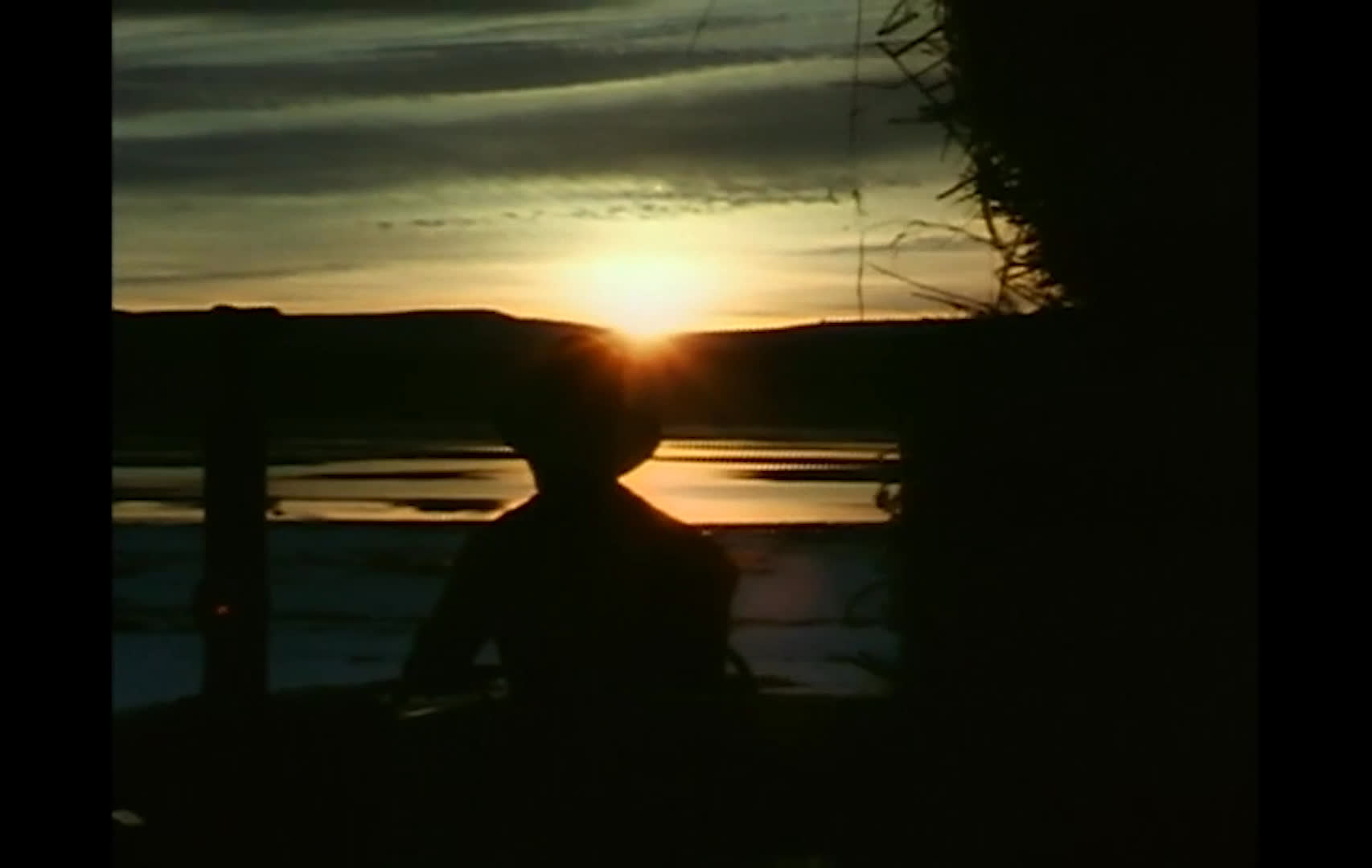 A hat-wearing farmer works atop a hay bale beneath a bright, cloud-filled sky, intercut with dark silhouettes against a glowing sunset over the rural landscape. The narration reflects on a farmer’s hope for a good year and the resilience to move past bad seasons.