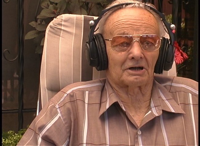 An elderly man wearing glasses and headphones sits in a chair while actively participating in a conversation. He speaks and listens, displaying engaged expressions throughout the interaction.