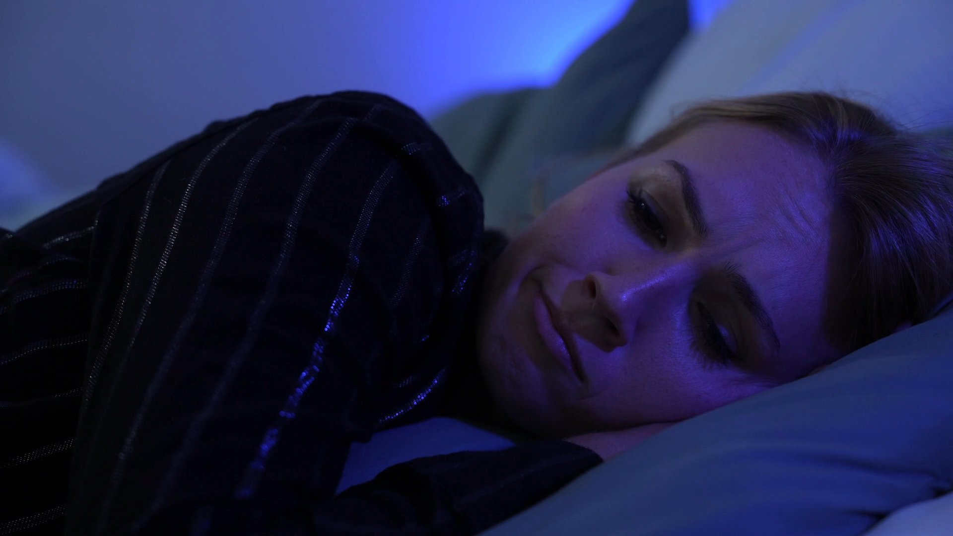 A tense interaction between a man and a woman in a living room shifts to a solitary, distressing night scene as the woman struggles with emotional turmoil while lying in bed.