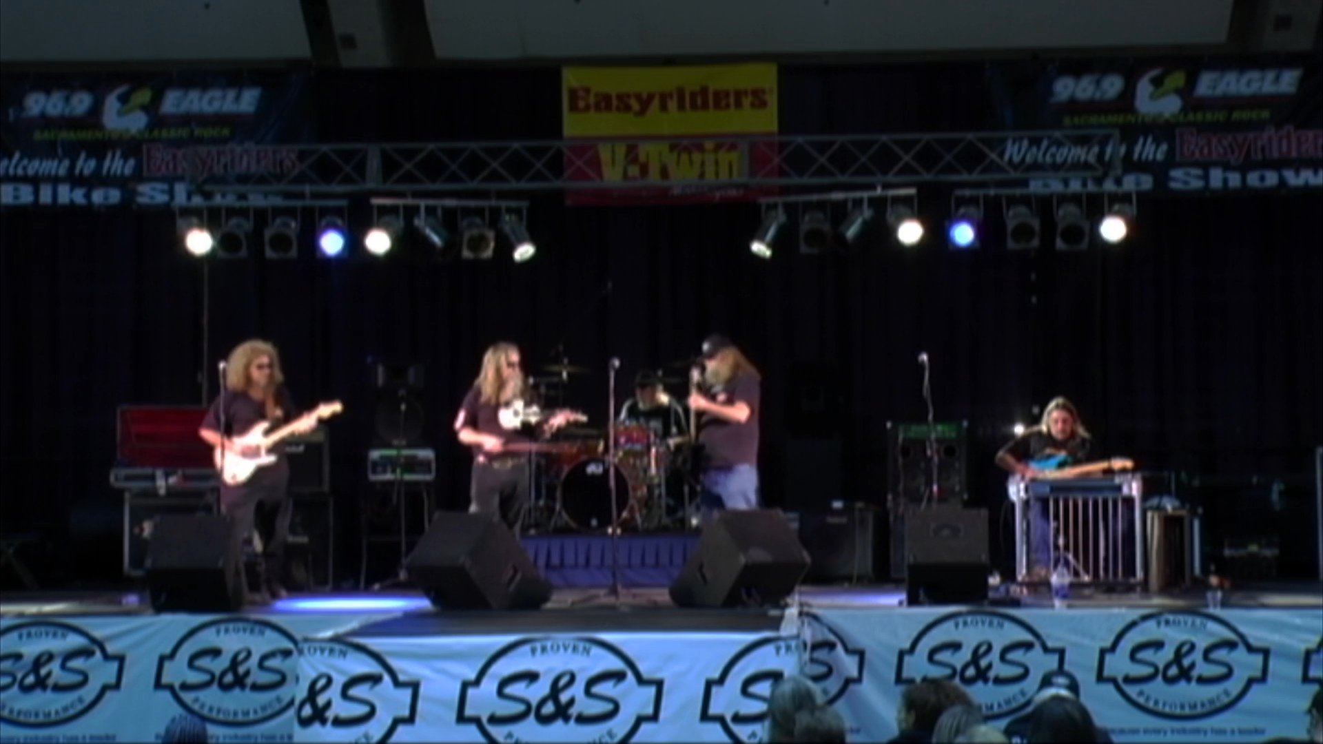 A musician plays an electric violin on a stage backed by a band performing live. The scene captures the ensemble during a concert performance at an event.