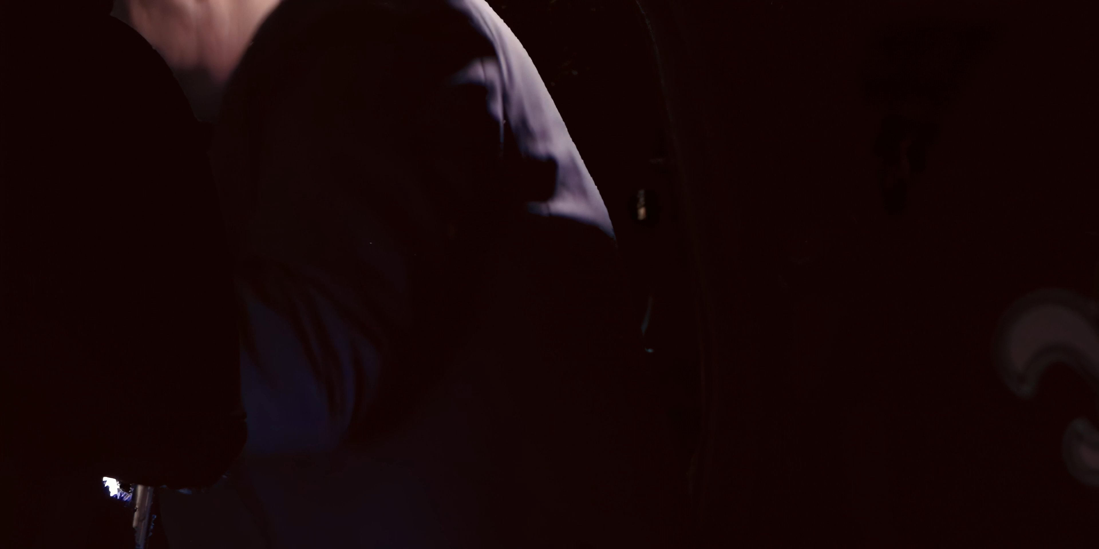 Two men engage in a tense conversation while seated inside a vehicle at night. The contrasting attire highlights the distinct personas of the individuals involved in the exchange.