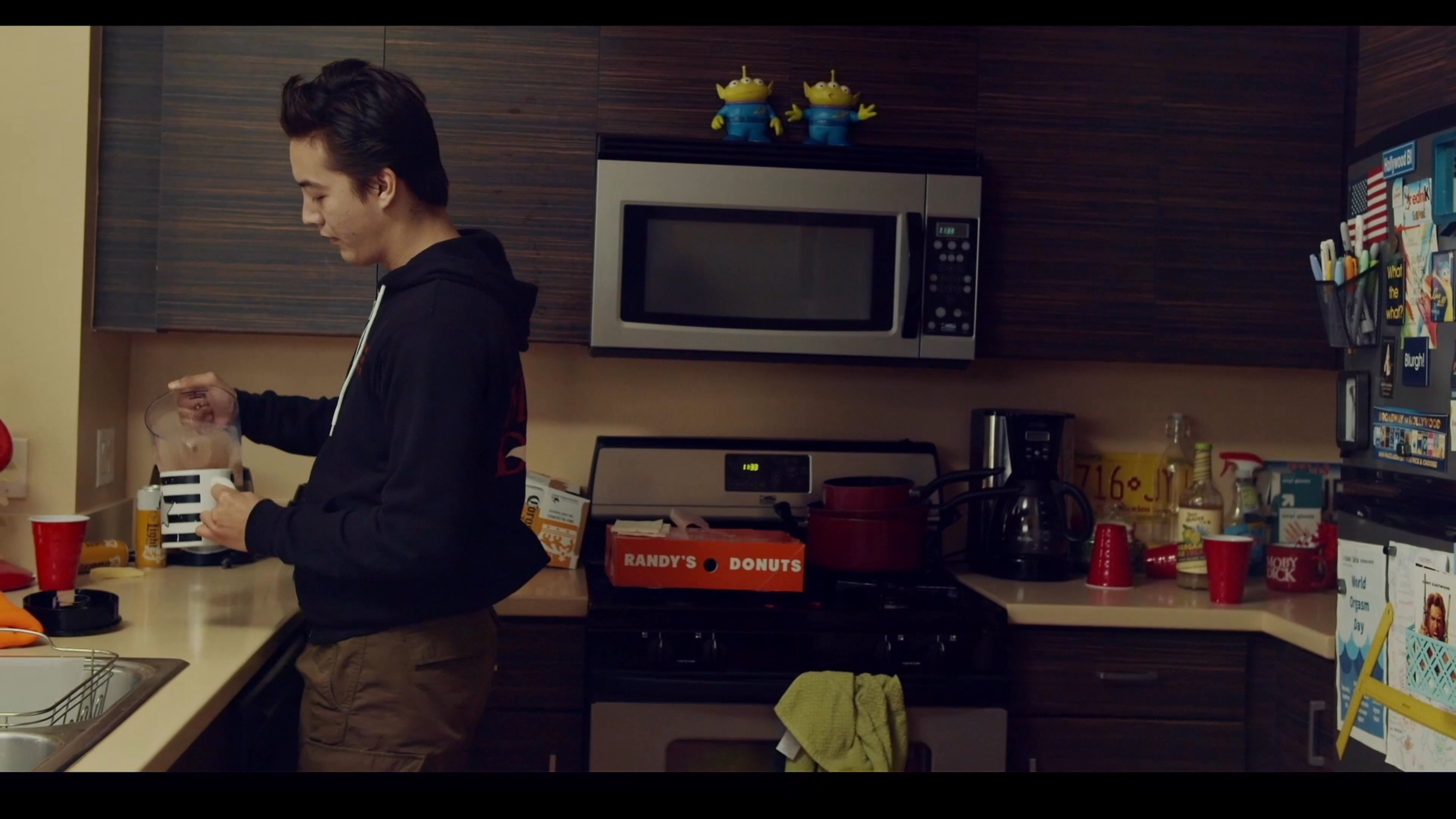 A young man transitions from reading about Stephen Hawking on his living room sofa to preparing and enjoying a hot beverage in his kitchen. The scene concludes with a focus on small collectible figurines placed on a table.