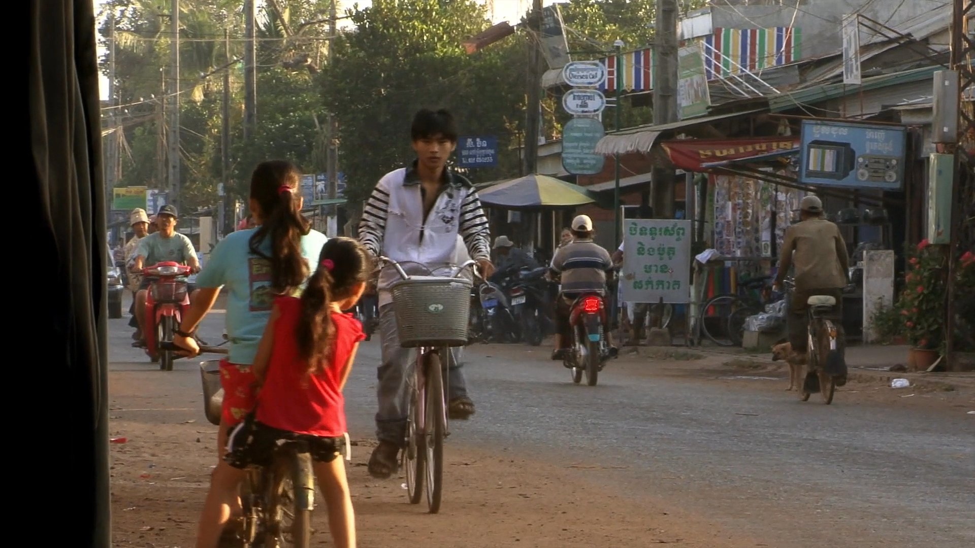 Daily life unfolds in a bustling Cambodian market street featuring commuters, traditional oxcart transport, and local vendors performing tasks like ice preparation. The sequence captures a vibrant cross-section of rural commerce and transit.