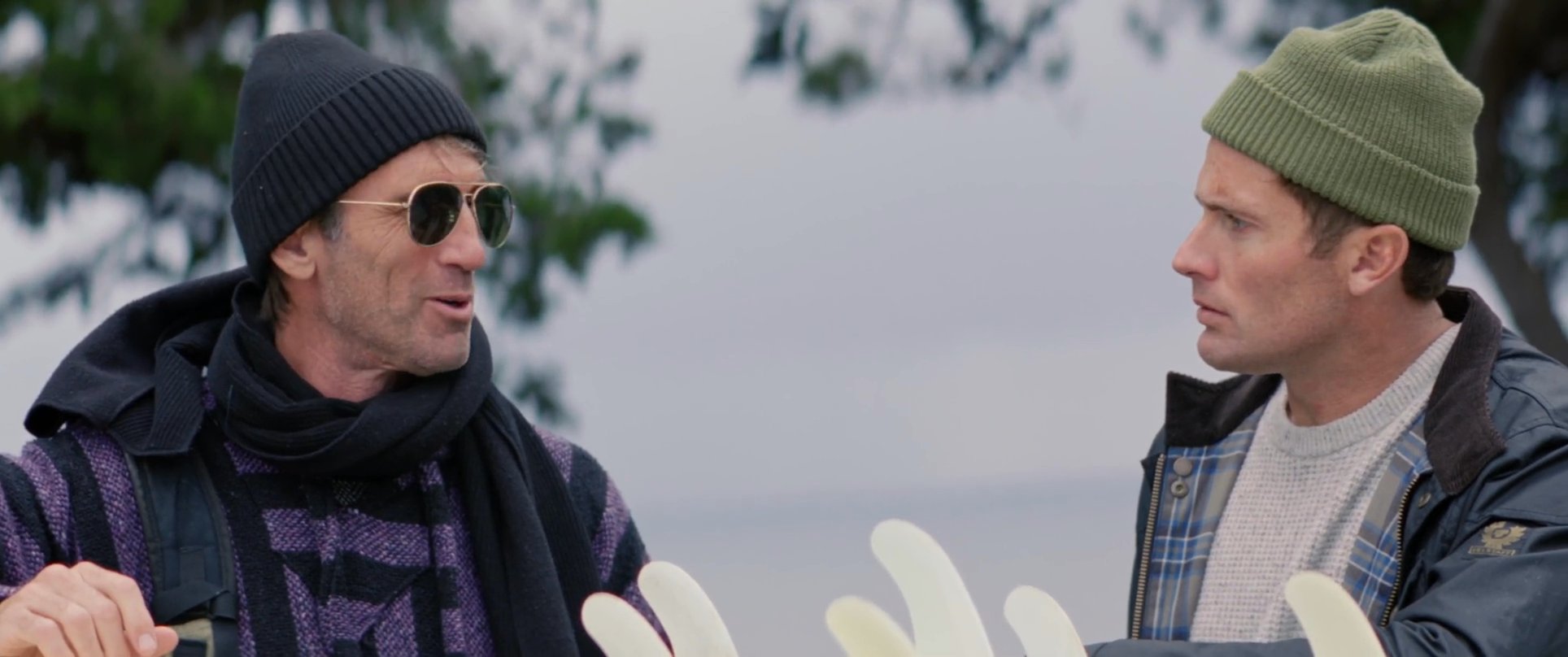 Three men wearing casual, layered outdoor clothing converse near the coast while preparing surfboards for an outing. The scene focuses on the interaction between the group as they handle their surf gear in an outdoor environment.