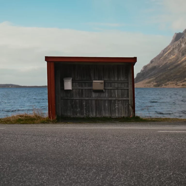 Video of man walking into frame to a small hut along the road against a lake pulling out a newspaper from a mailbox