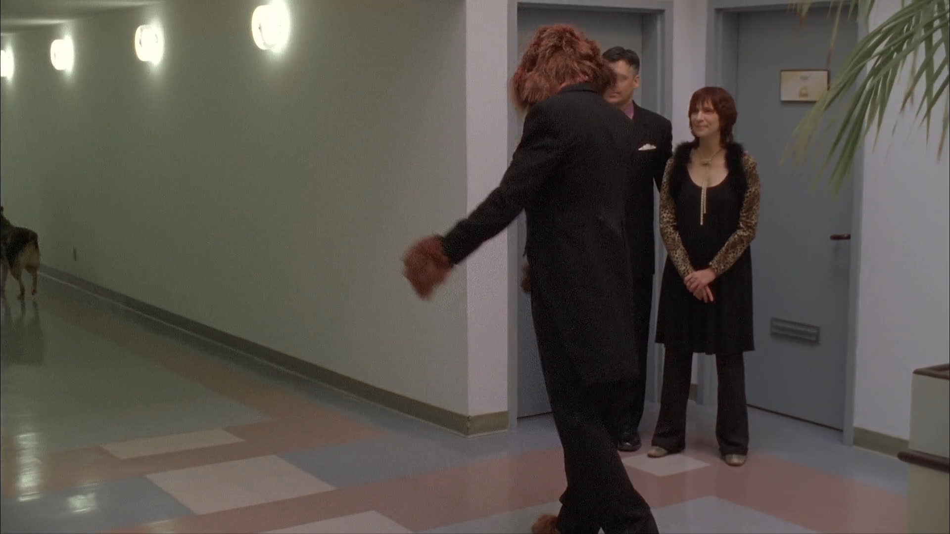 A man wearing a fur-collared coat walks a dog through a residential hallway while two formally dressed individuals observe. The man and his dog depart, leaving the two observers standing alone in the corridor.