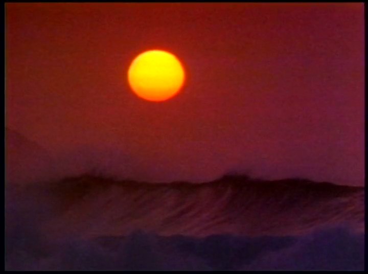 A surfer enters the ocean with a red longboard before transitioning to paddling out as the sun sets over the water. The scene captures the transition from daylight preparation to a serene evening session on the water.