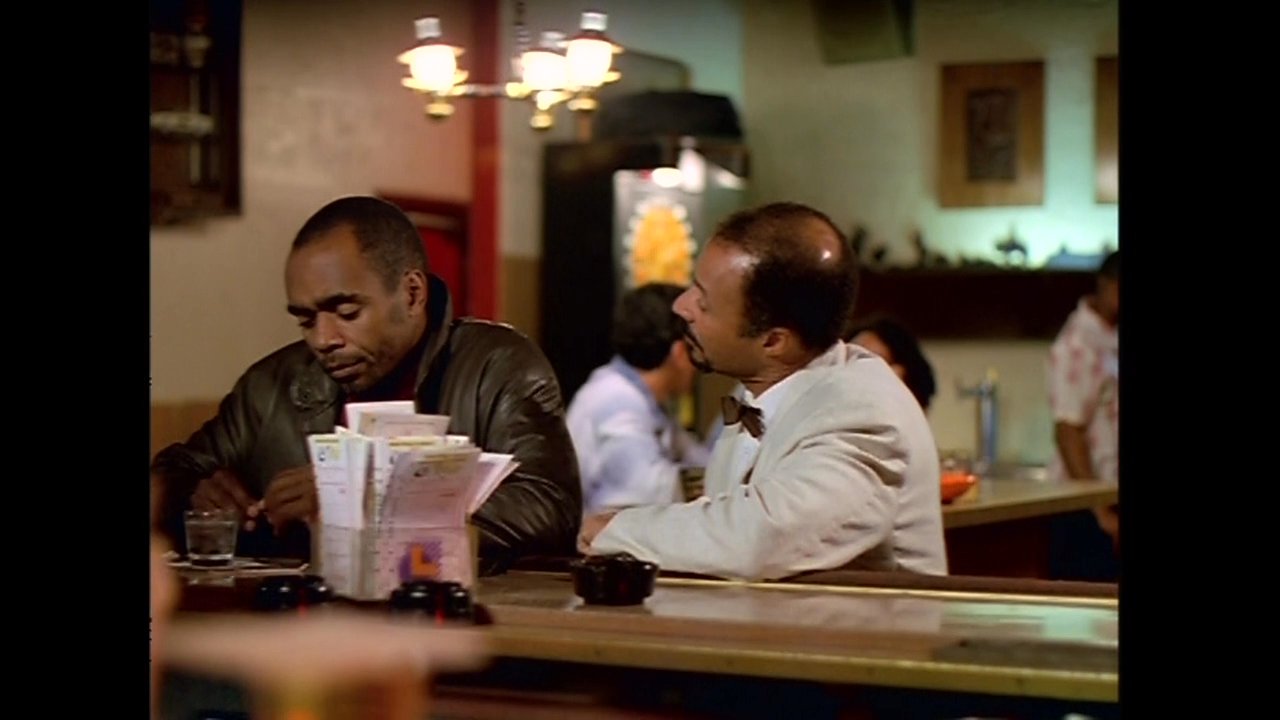Two men sit at a dimly lit bar, engaged in an earnest, private conversation amidst a social setting.
