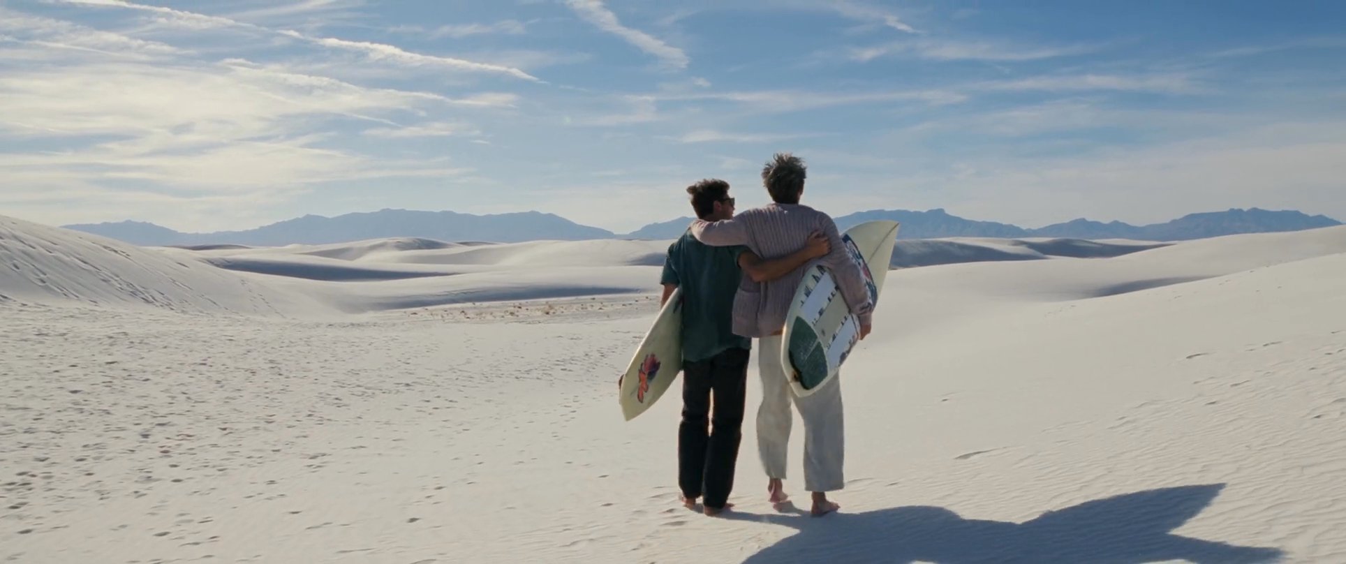 Two individuals carrying surfboards walk arm-in-arm across an expansive landscape of bright white sand dunes under a clear blue sky.