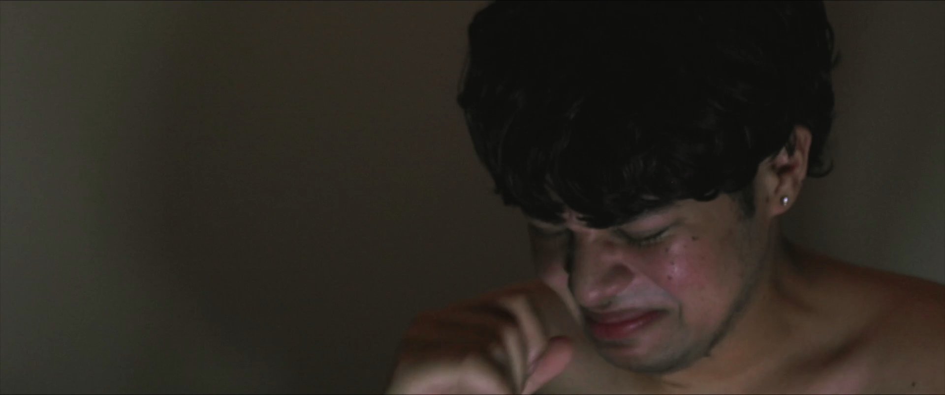 A young man experiences a moment of intense emotional vulnerability while lying in bed and reflecting in a dimly lit room. The sequence captures his raw expression of grief and internalized pain.