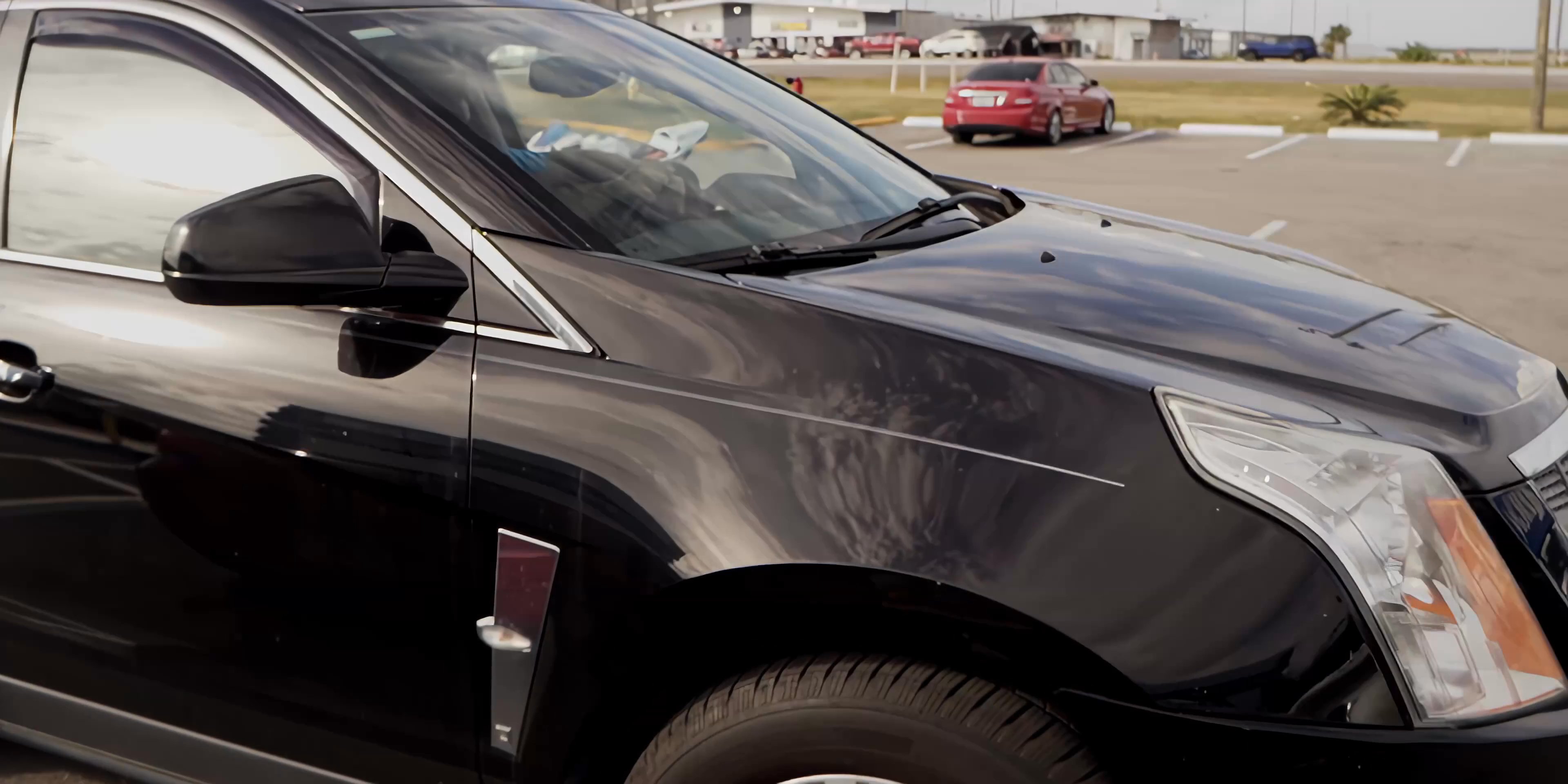A man receives an item through a car window before the vehicle drives away across a parking lot toward a building entrance.
