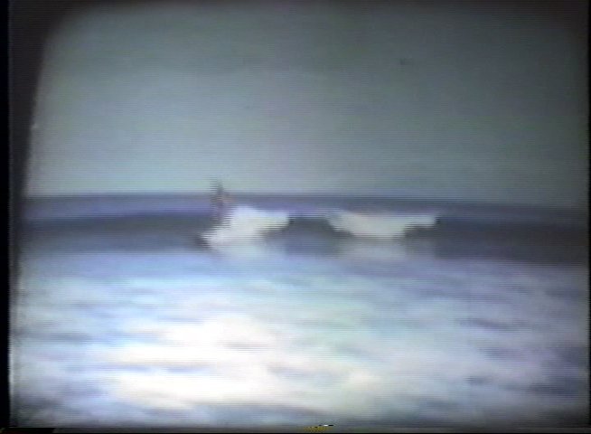 A surfer maneuvers along a wave near a pier in this vintage film footage. The sequence captures the progression of the ride from initial catch to the surfer exiting the wave.