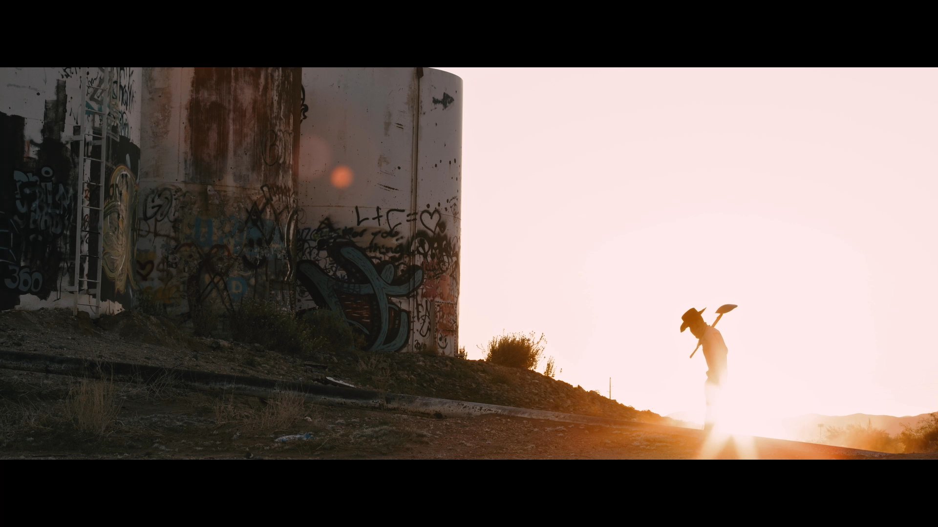 A laborer walks through a desert landscape at sunset carrying a shovel, underscoring themes of migration and struggle. The scene introduces the documentary title 'Entre Fronteras' set against a stark, backlit backdrop.